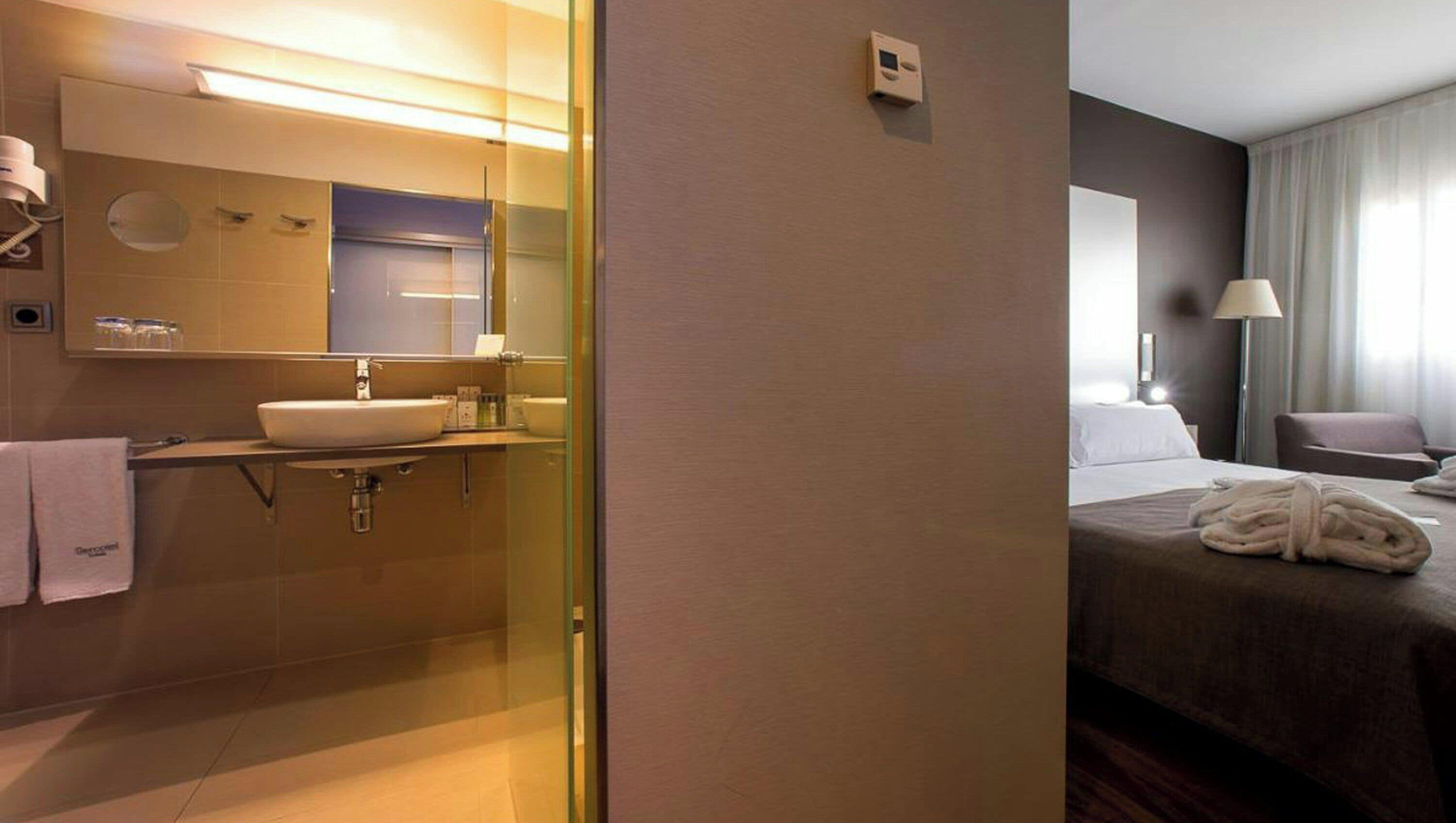 A hotel room with a bathroom featuring a sink, mirror, and towel rack. The room has a bed with a blanket and a bedside table with a lamp.