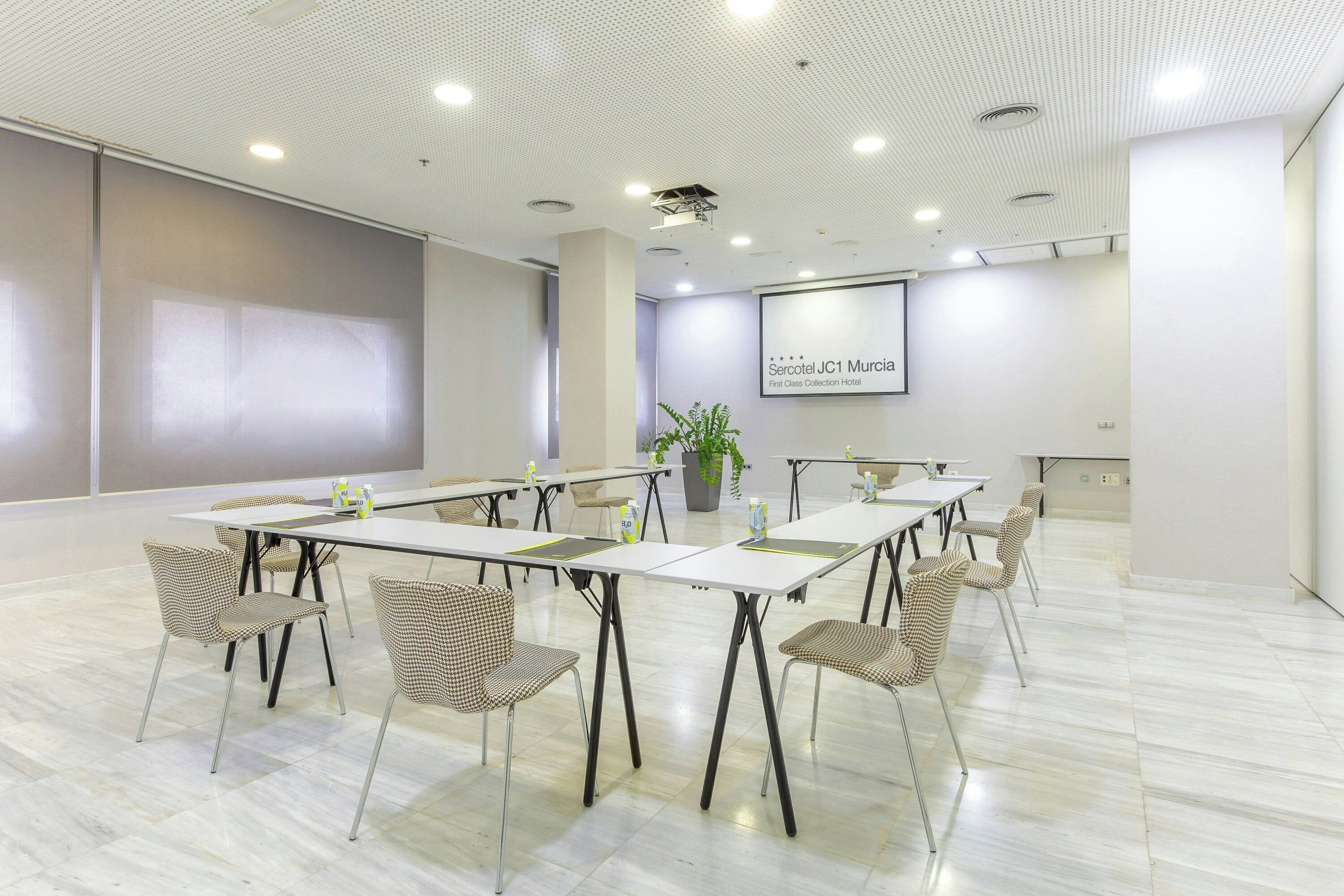 Un salón de conferencias con una pantalla grande, mesas blancas y sillas de estampado pata de gallo.