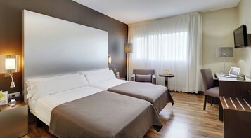 A hotel room with two beds, a desk with a laptop, a chair, a small table with a tray, and a TV mounted on the wall.