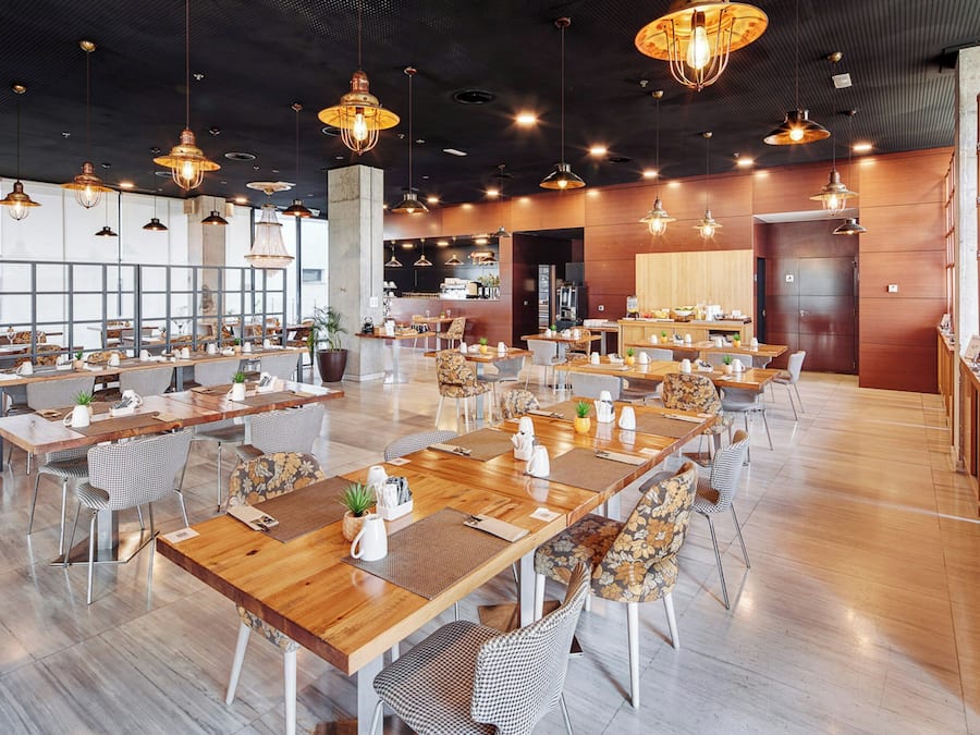 A spacious restaurant with multiple dining tables and chairs, large windows, and hanging pendant lights.