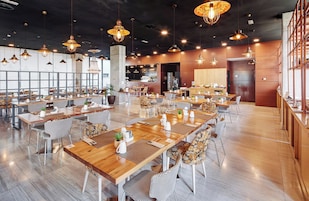 A spacious restaurant with multiple dining tables and chairs, large windows, and hanging pendant lights.