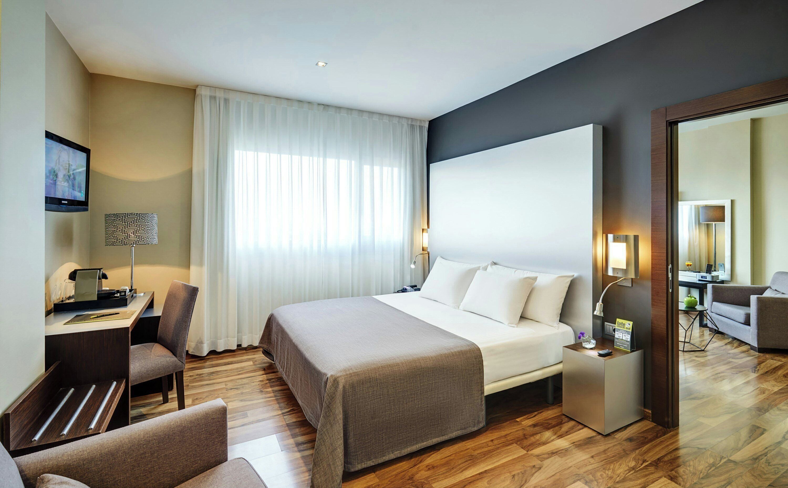 Una habitación de hotel con cama, mesitas de noche, escritorio, silla, televisor y un espejo grande.