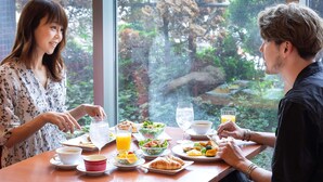 Een eettafel met diverse ontbijtartikelen en dranken, met op de achtergrond een groot raam met uitzicht op bomen en een gebouw.