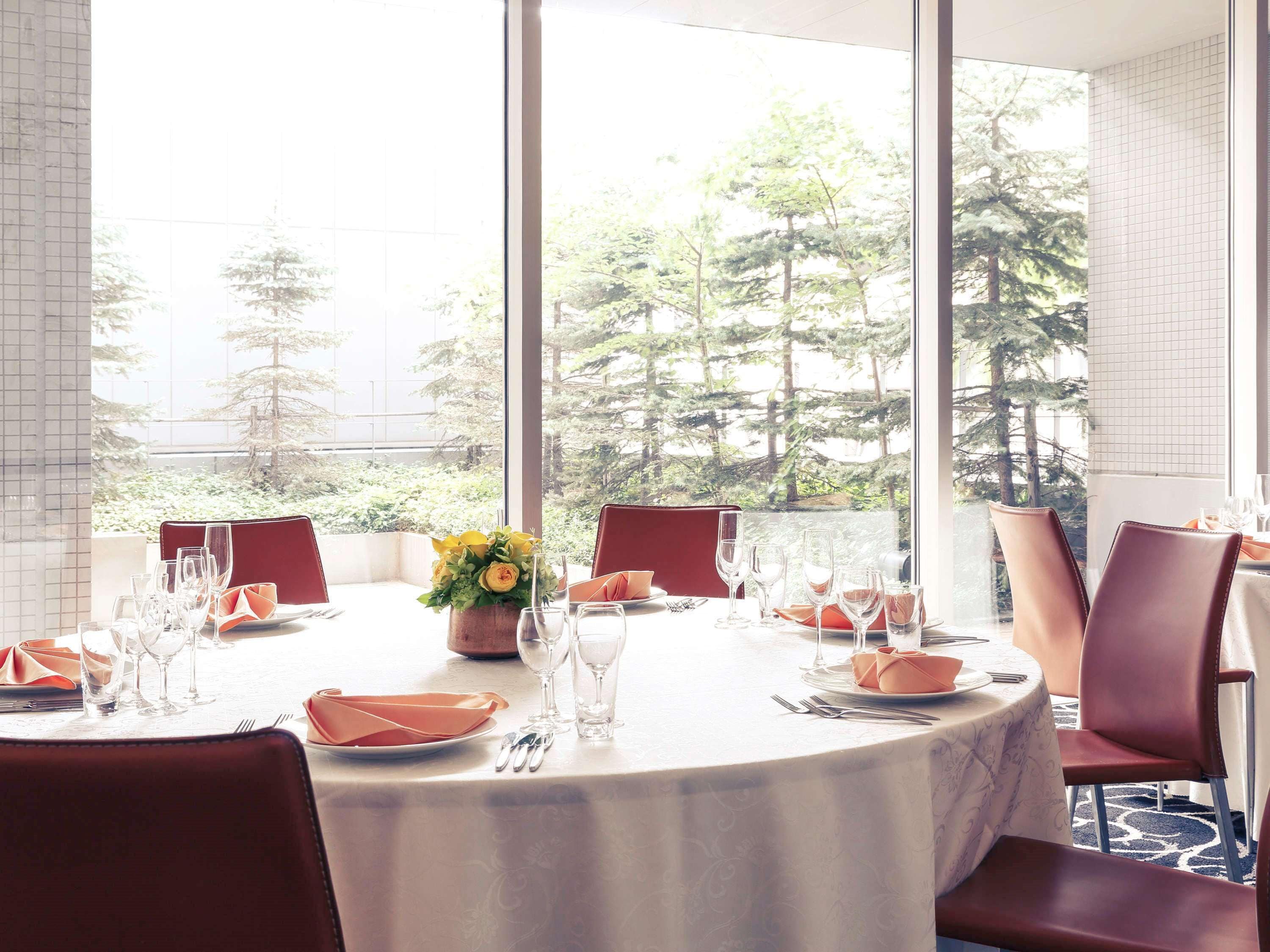A dining area with round tables, red chairs, and a view of trees through large windows.