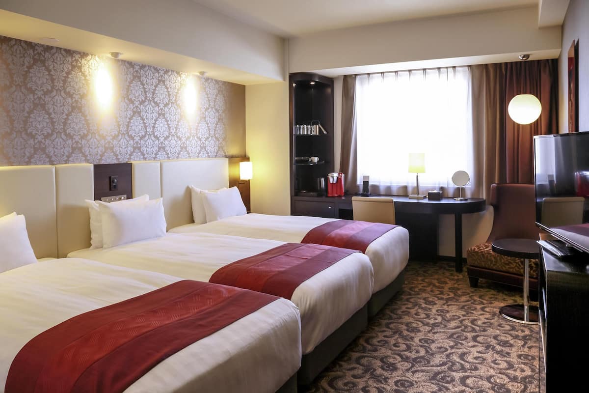 A hotel room with two beds, a desk, a TV, and a window with curtains.