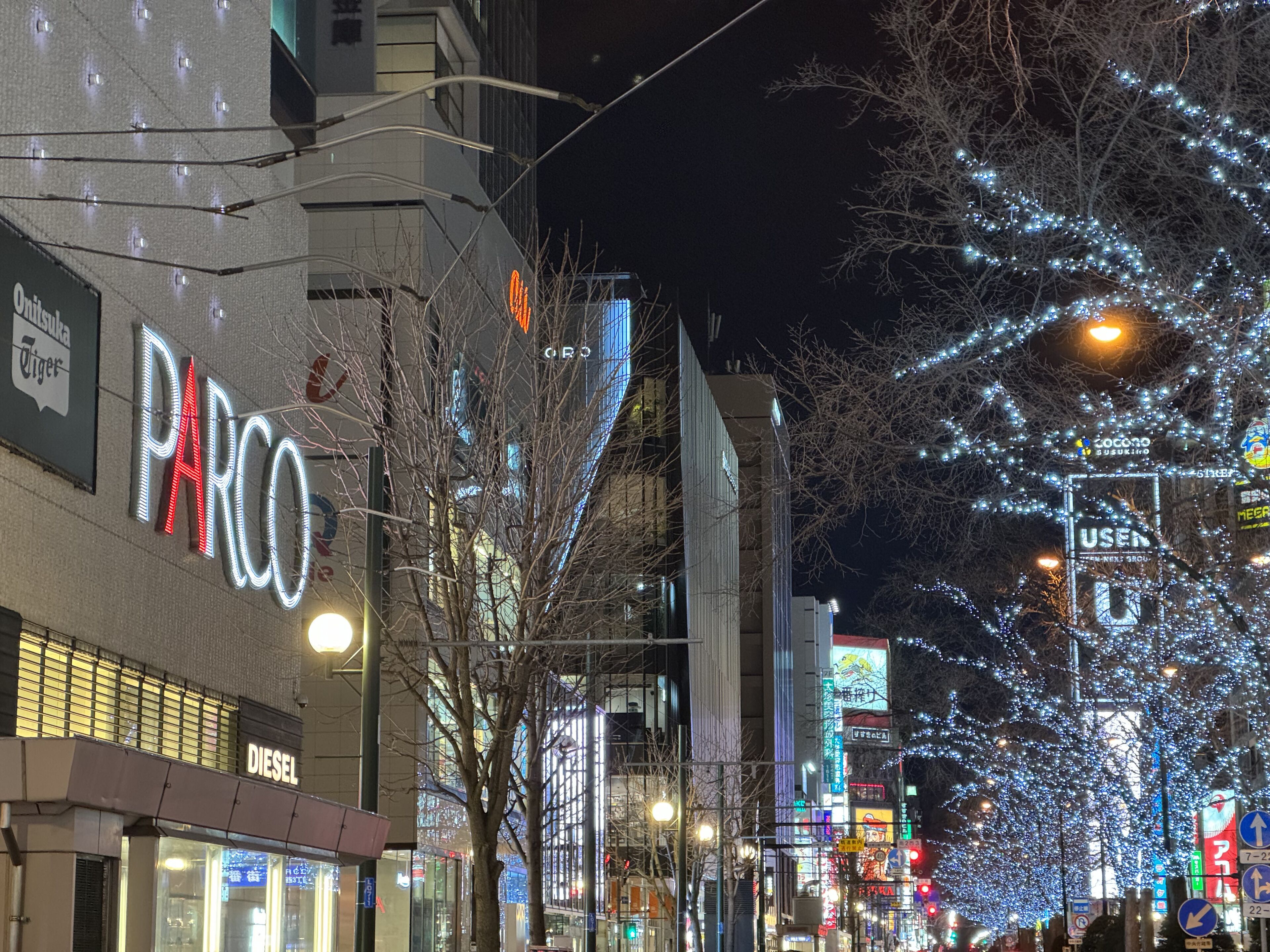 Una calle urbana de noche con escaparates iluminados, incluyendo una tienda PARCO y una tienda DIESEL.
