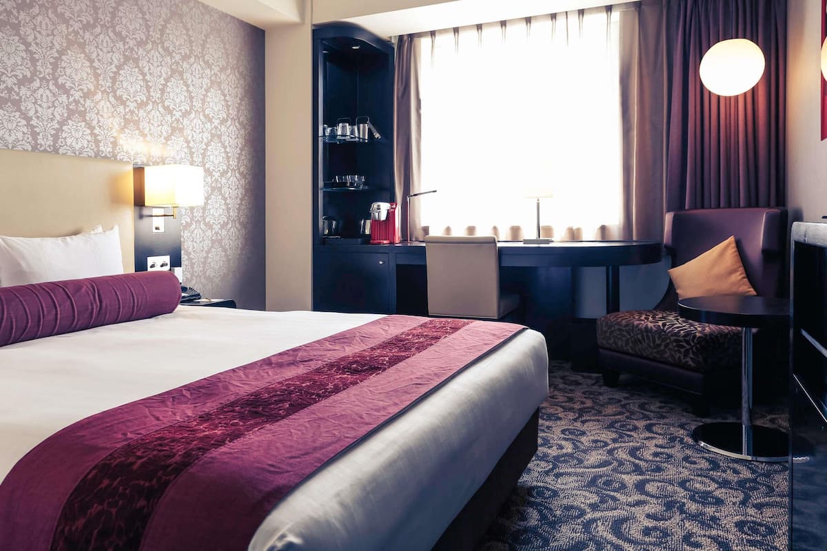 A hotel room with a large bed, a desk with a chair, a minibar, and a view of the outside.