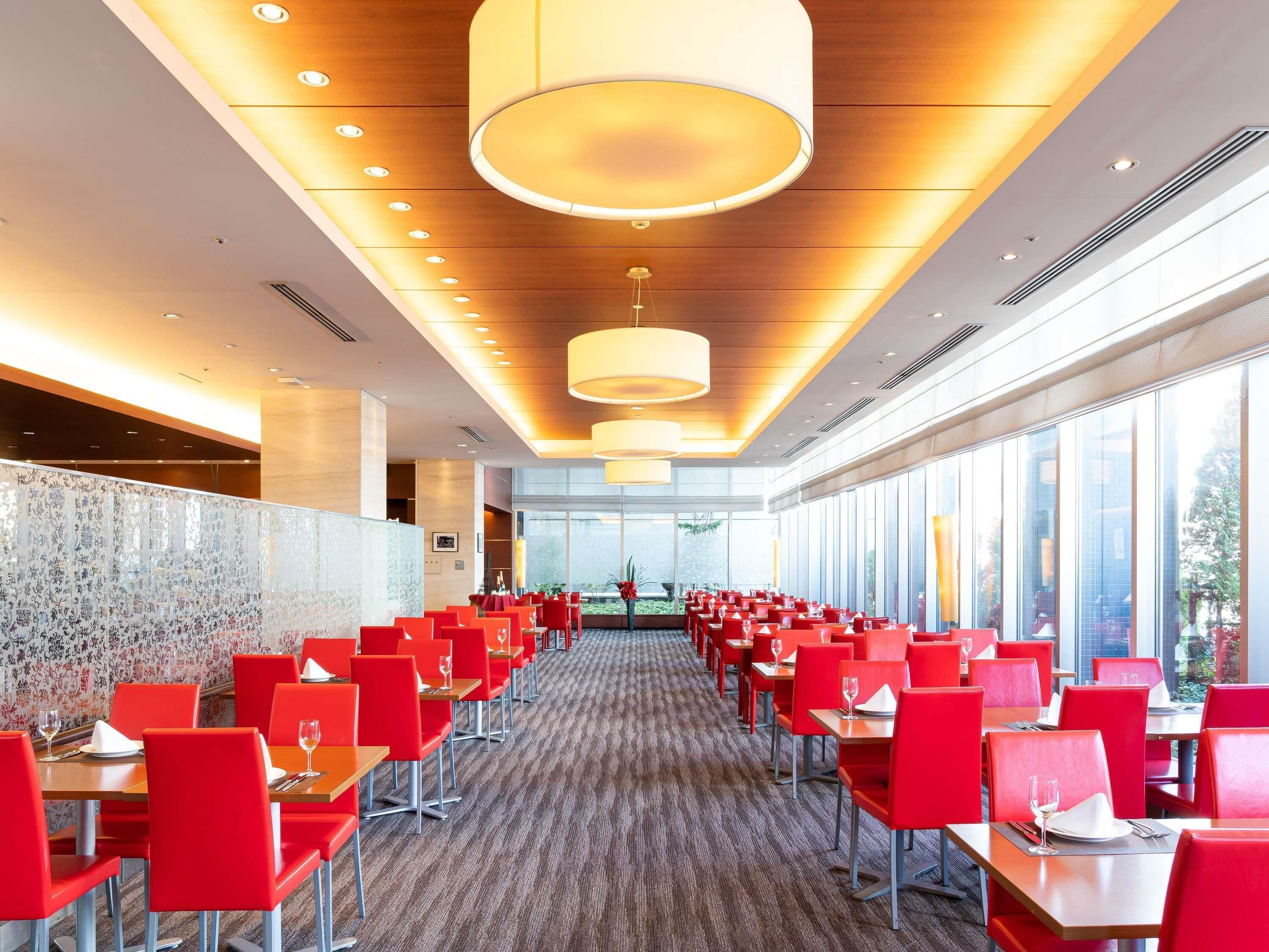 A spacious dining area with red chairs and tables, large windows, and modern pendant lighting.