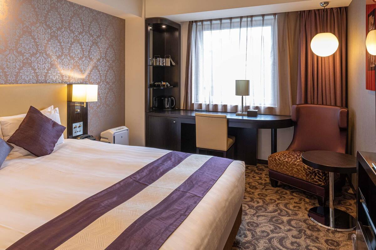 A hotel room with a bed, desk, chair, and a view of the city.