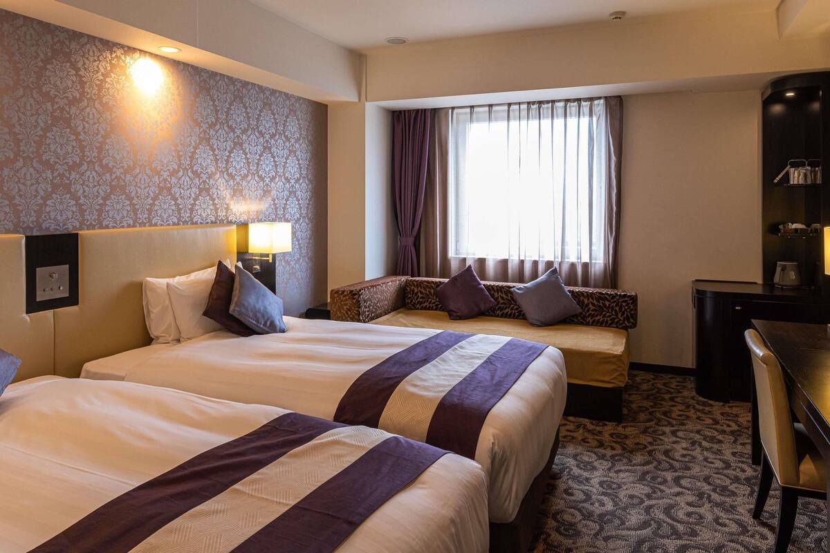 A hotel room with two beds, a desk, a chair, a window with curtains, and a patterned carpet.