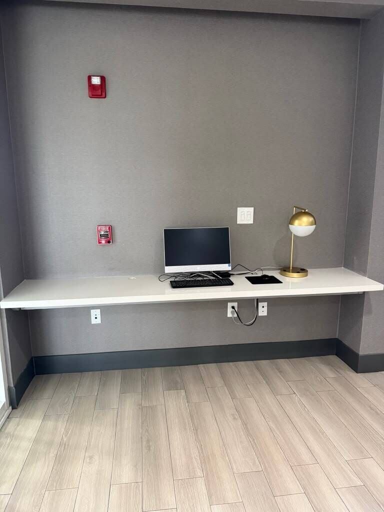 A room with a white desk, a laptop, a gold desk lamp, and a fire alarm on the wall.