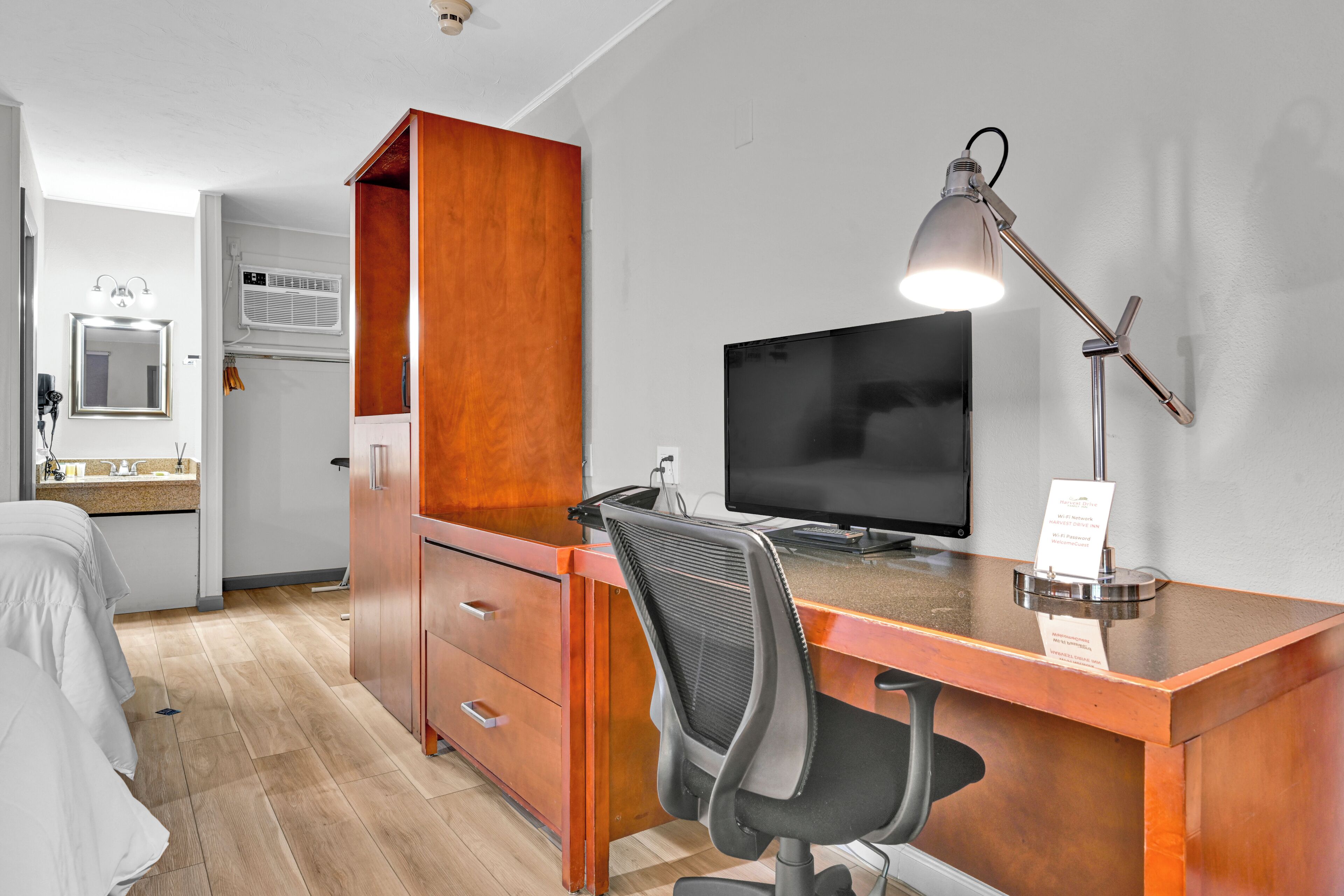 A hotel room with a wooden desk, a computer, a desk lamp, a TV, a bed, and a bathroom with a mirror and sink.