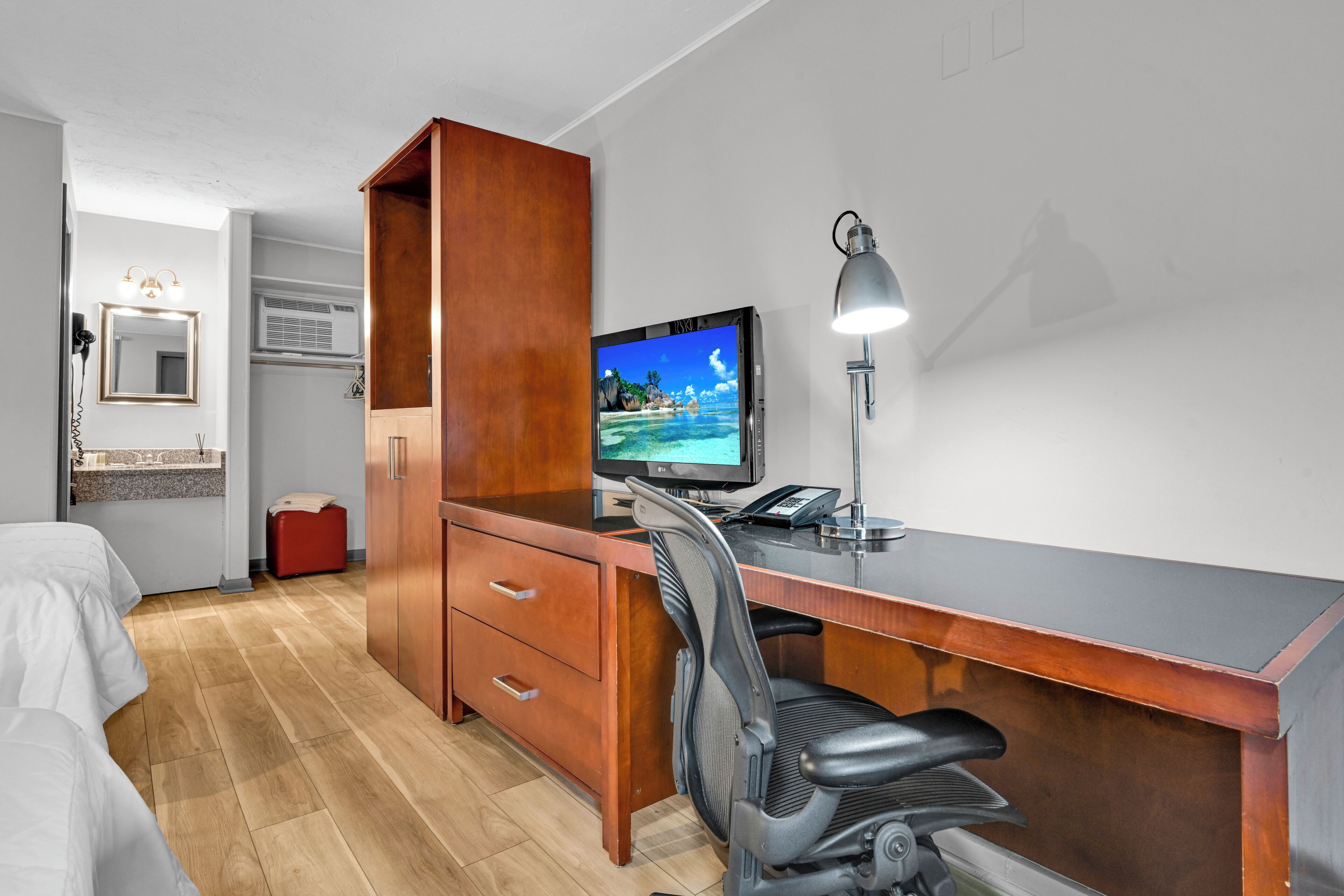 A hotel room with a wooden desk, a computer, a chair, a bed, a mirror, and a window with a view of a beach.
