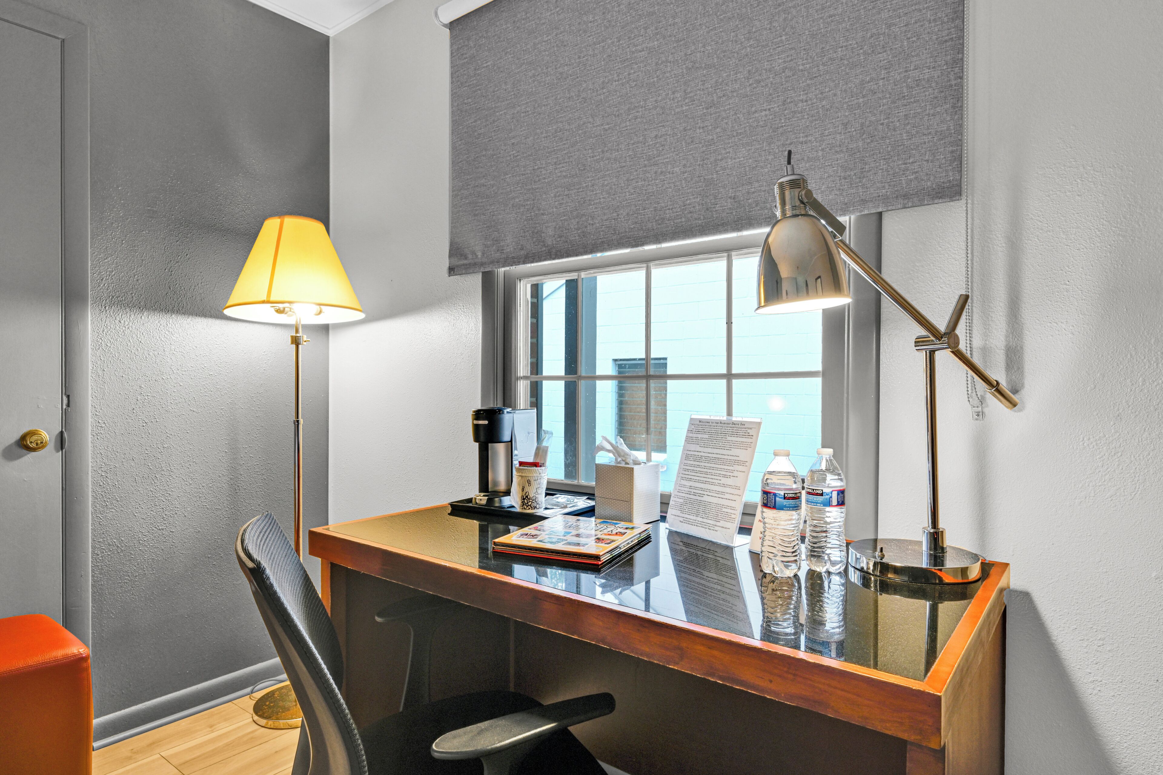 A hotel room with a wooden desk, two lamps, a chair, a window with blinds, a door, and a small trash bin.