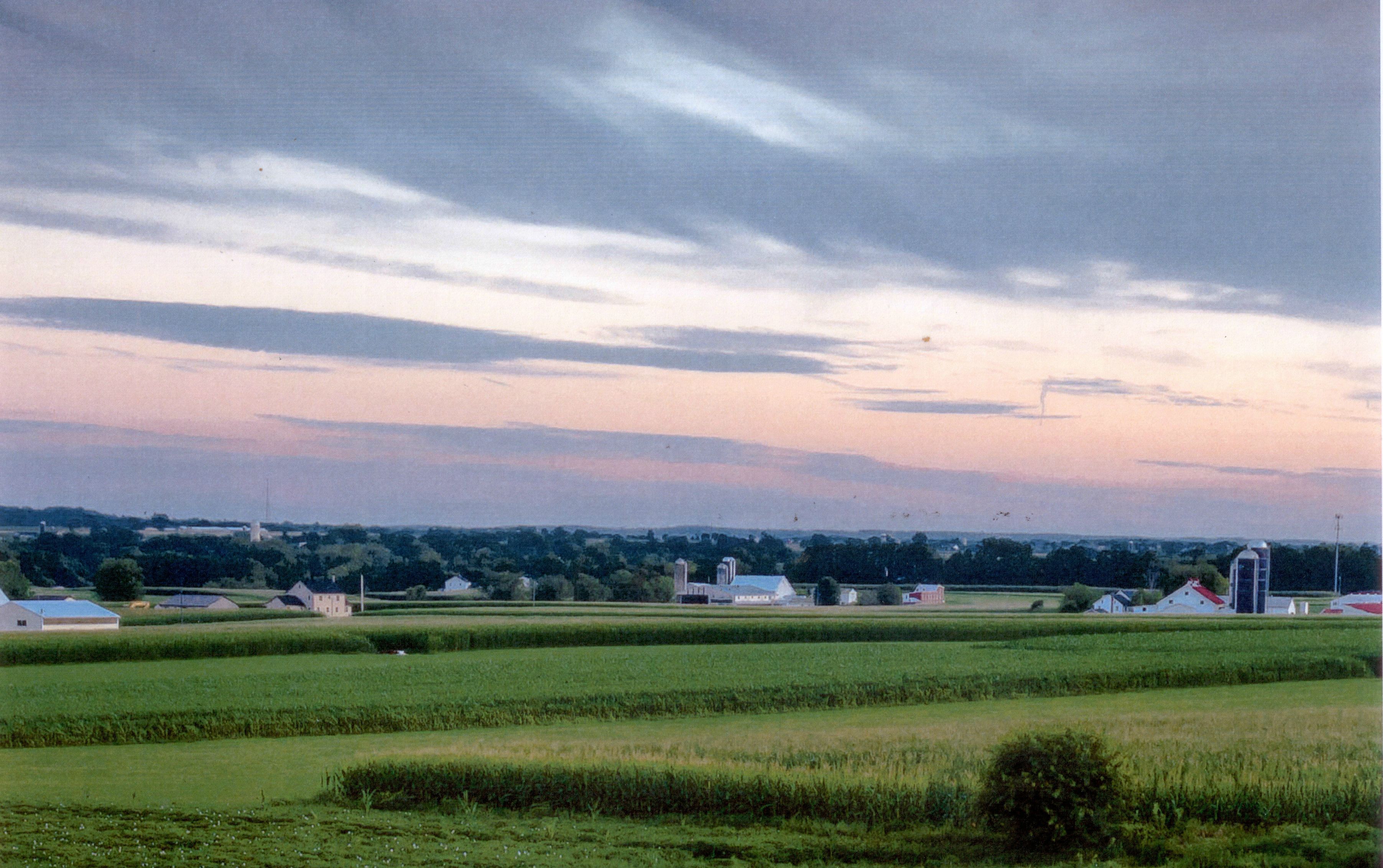Een landelijk landschap met velden, schuren en enkele huizen onder een bewolkte lucht.