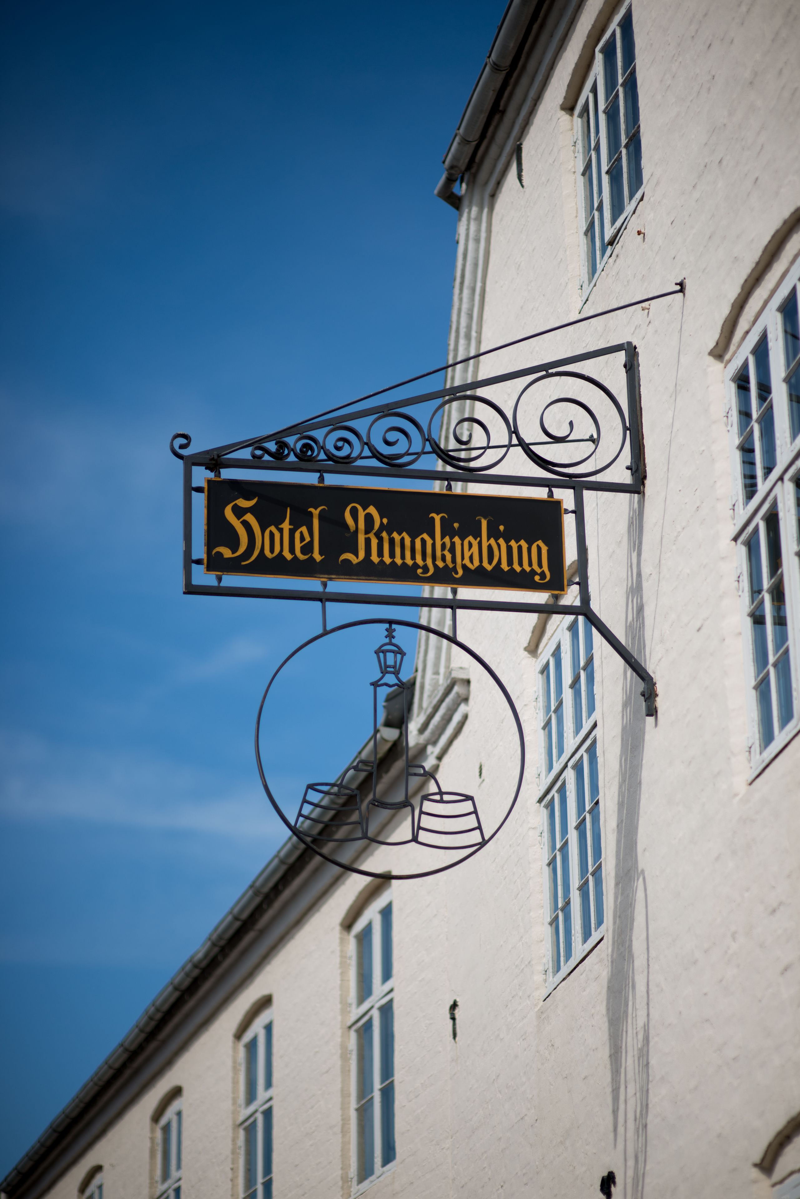 A sign for Hotel Ringkobing with a classic design, including a lamp and a bell, mounted on a building with multiple windows.