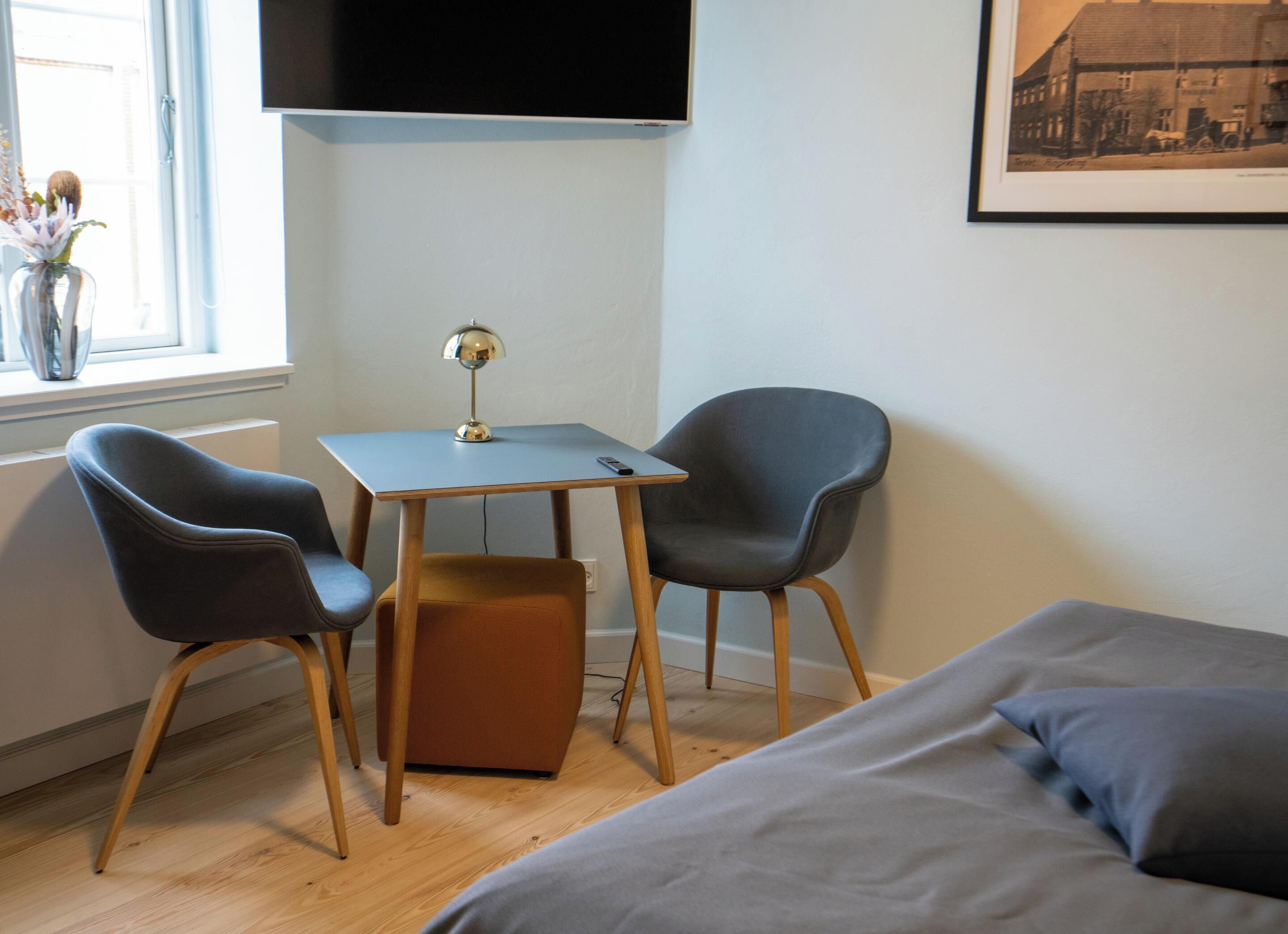 Una habitación con una cama, dos sillas, una mesita, una lámpara y un televisor en la pared.