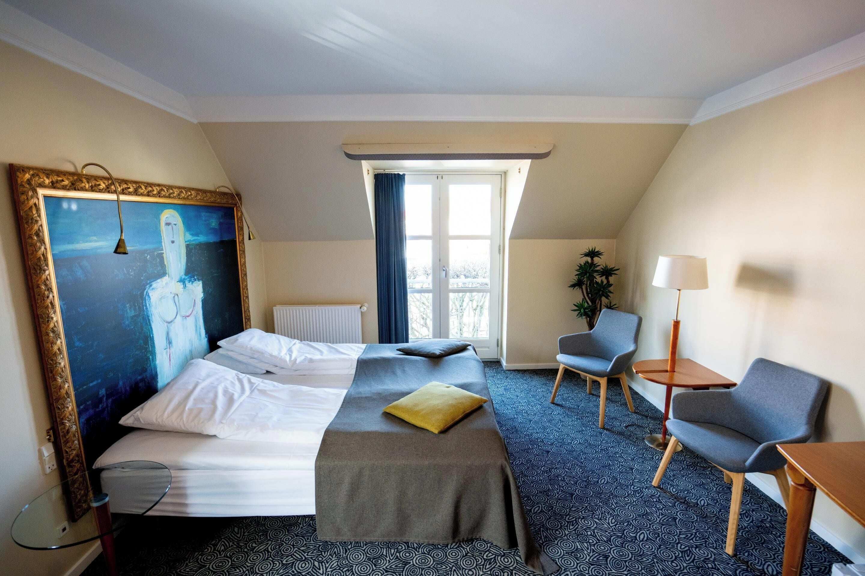 Habitación de hotel con cama, dos sillas, una mesa, un cuadro y ventana con cortinas.