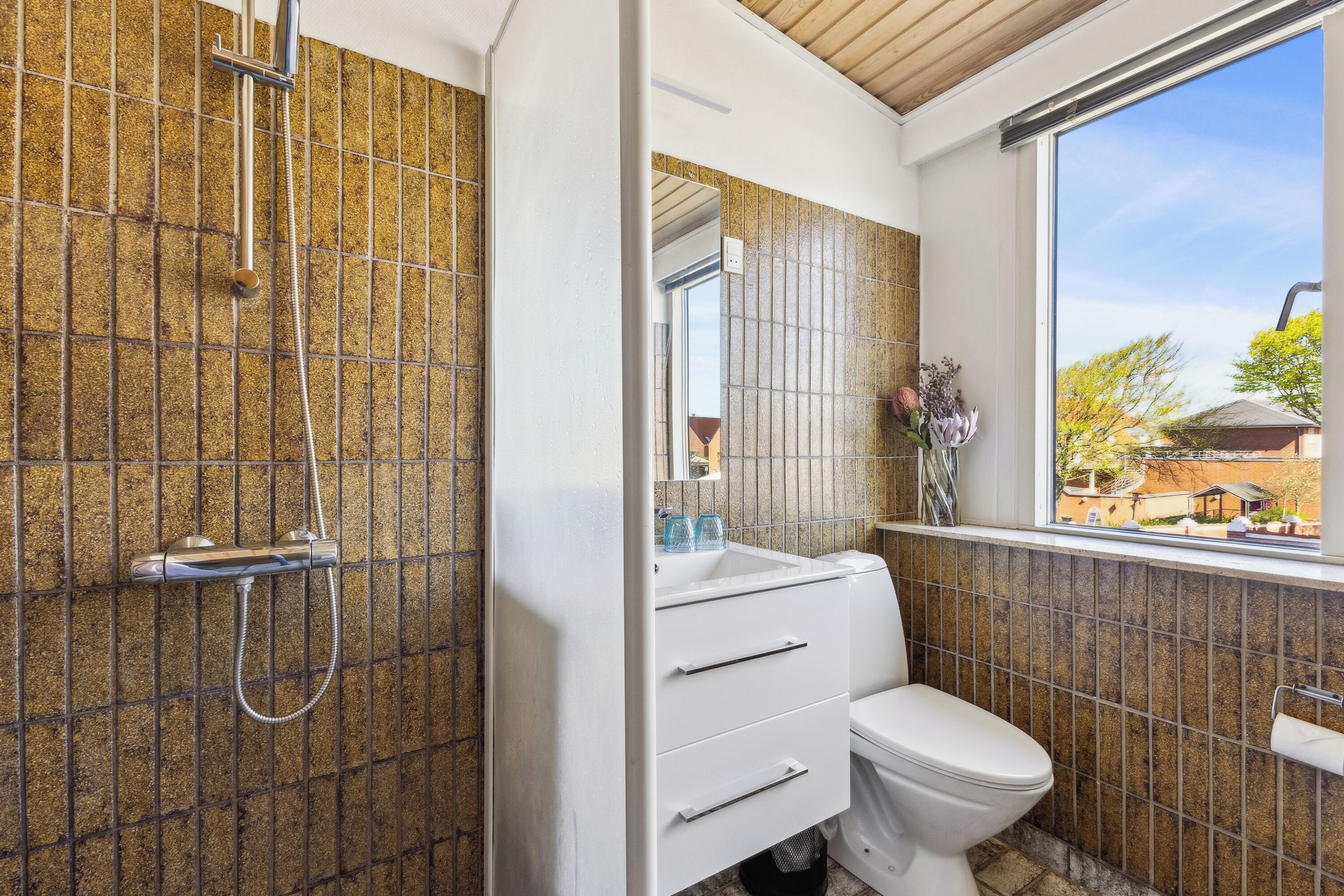 A bathroom with a shower, toilet, and sink, featuring a tiled wall and a window with a view.