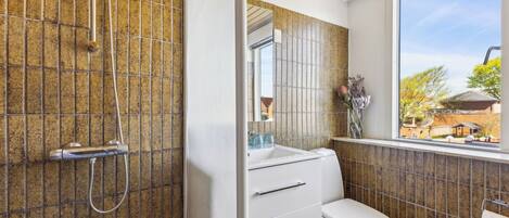 A bathroom with a shower, toilet, and sink, featuring a tiled wall and a window with a view.