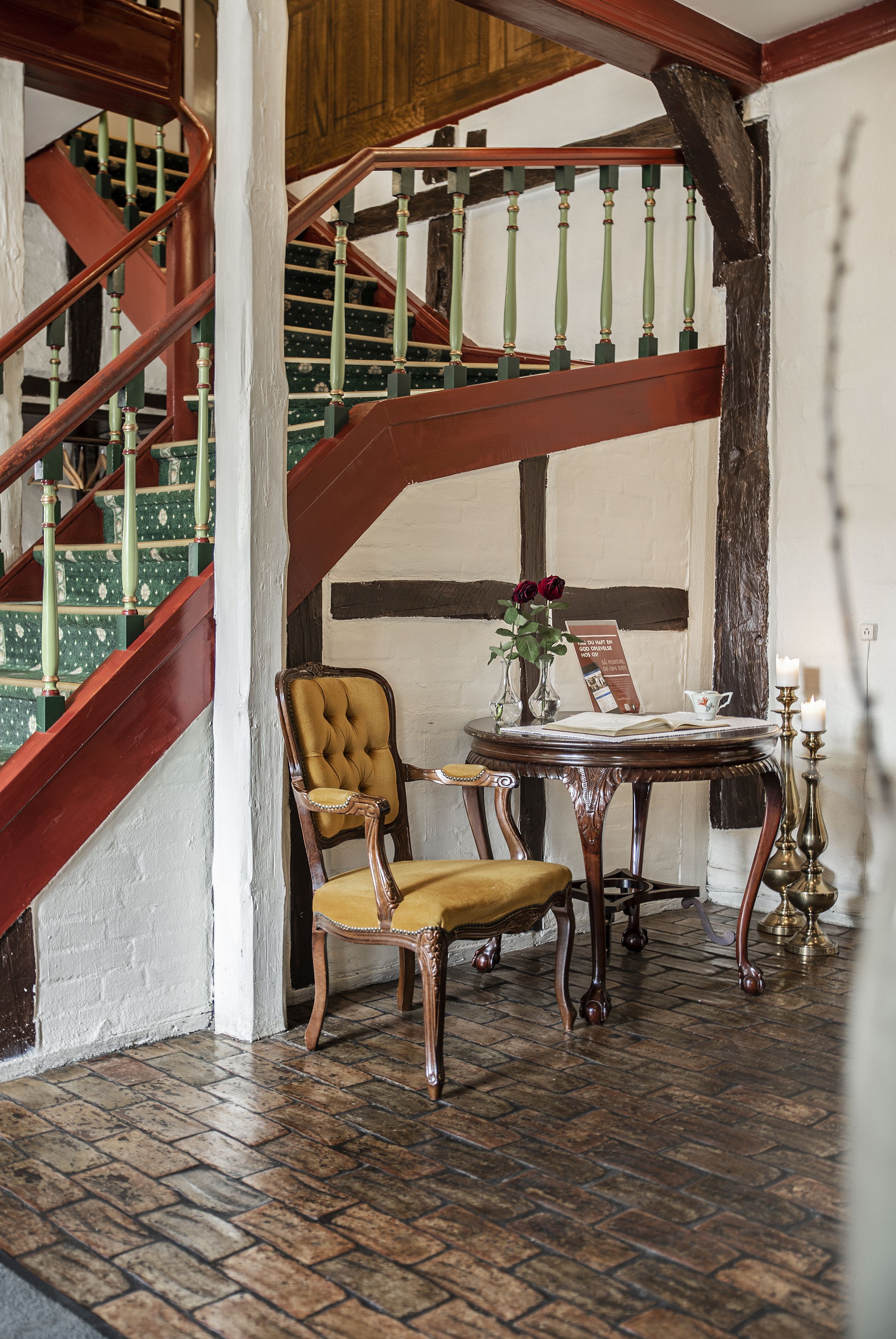 Un escalier avec des balustrades vertes, une chaise en bois, une table avec un livre et un vase de fleurs, et un chandelier.