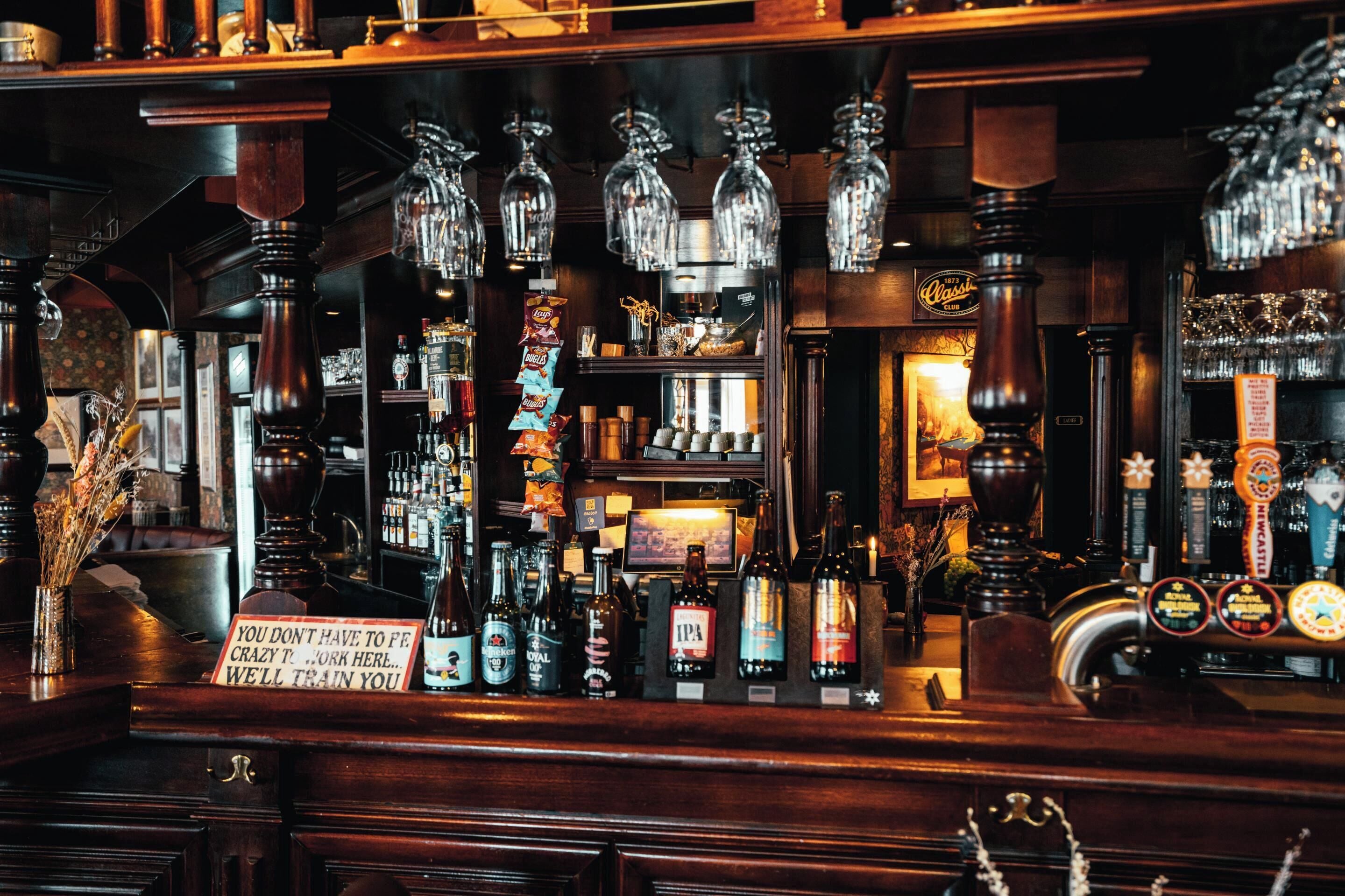 Un bar con varias botellas, vasos y un letrero que dice 'No hace falta estar loco para trabajar aquí... te entrenaremos'.