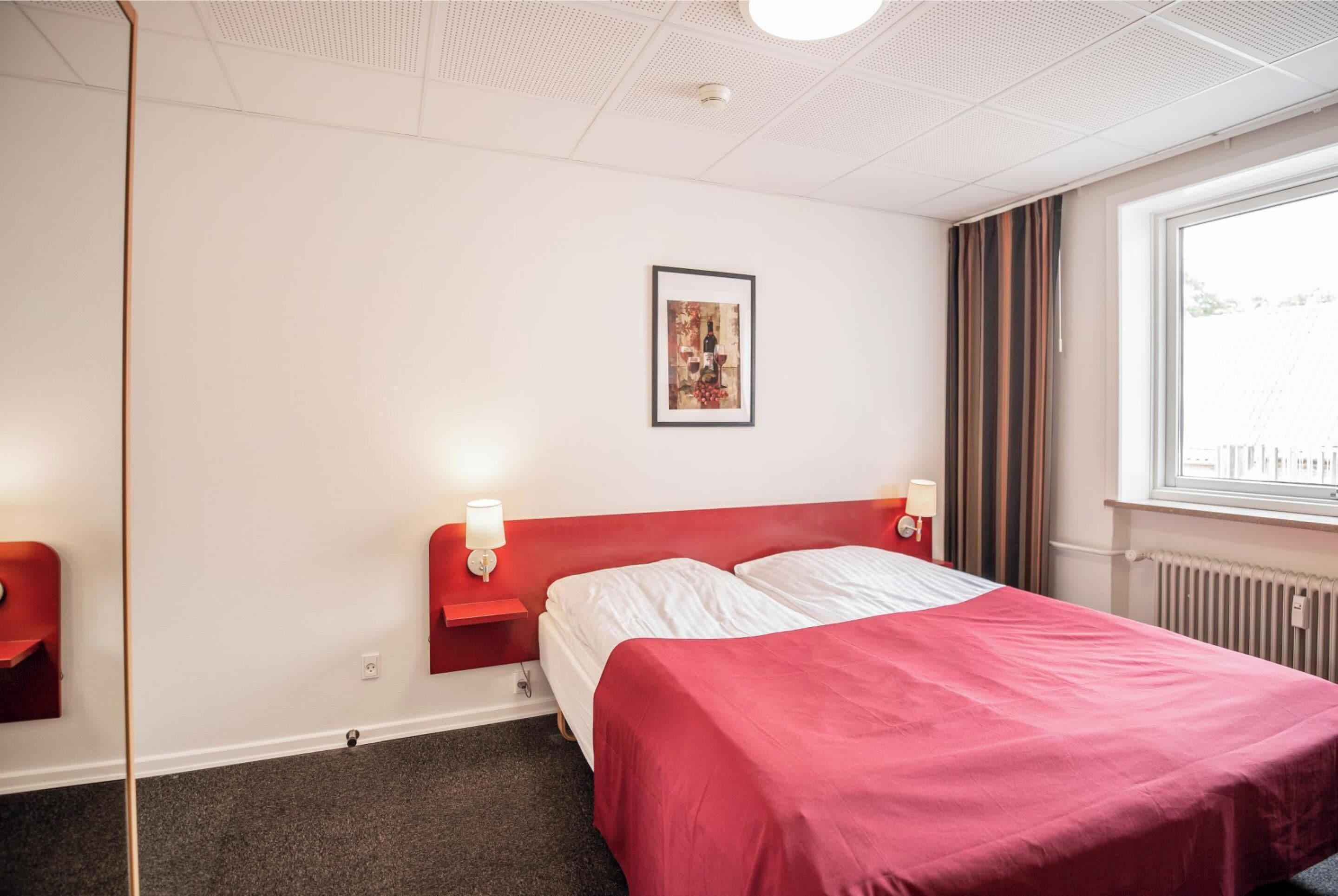 A hotel room with a red bedspread, two bedside tables with lamps, a framed picture on the wall, and a window with curtains.