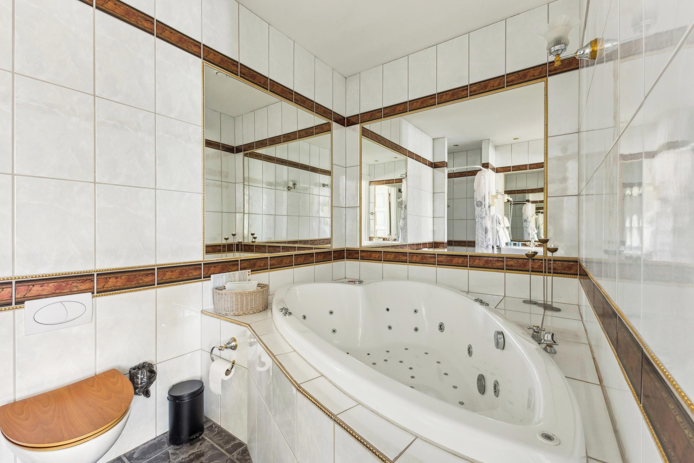 A bathroom with a large bathtub, a wooden toilet seat, a trash bin, and a mirror with a wooden frame.