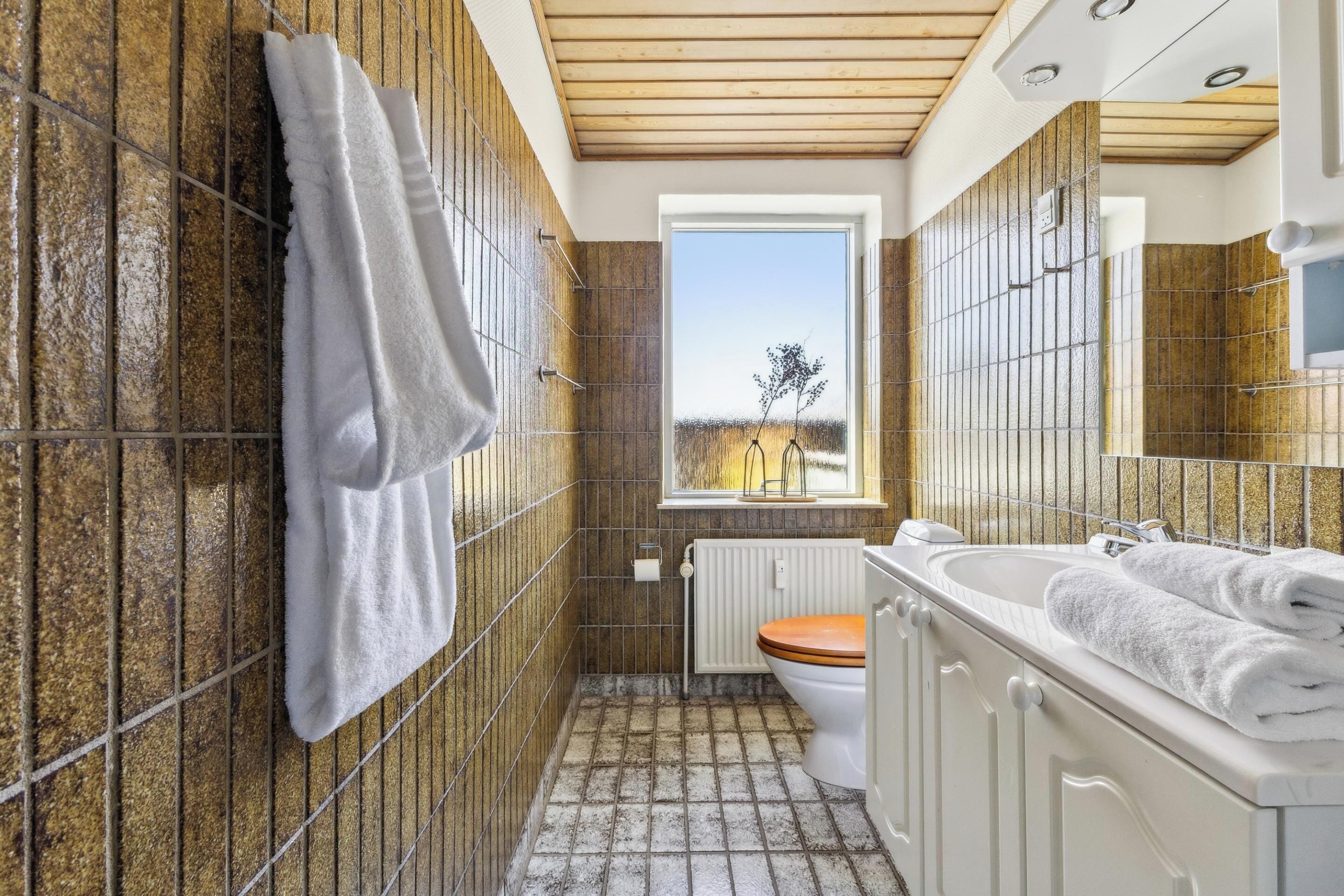 A bathroom with a wooden ceiling, a window with a view, a white sink, and a toilet.