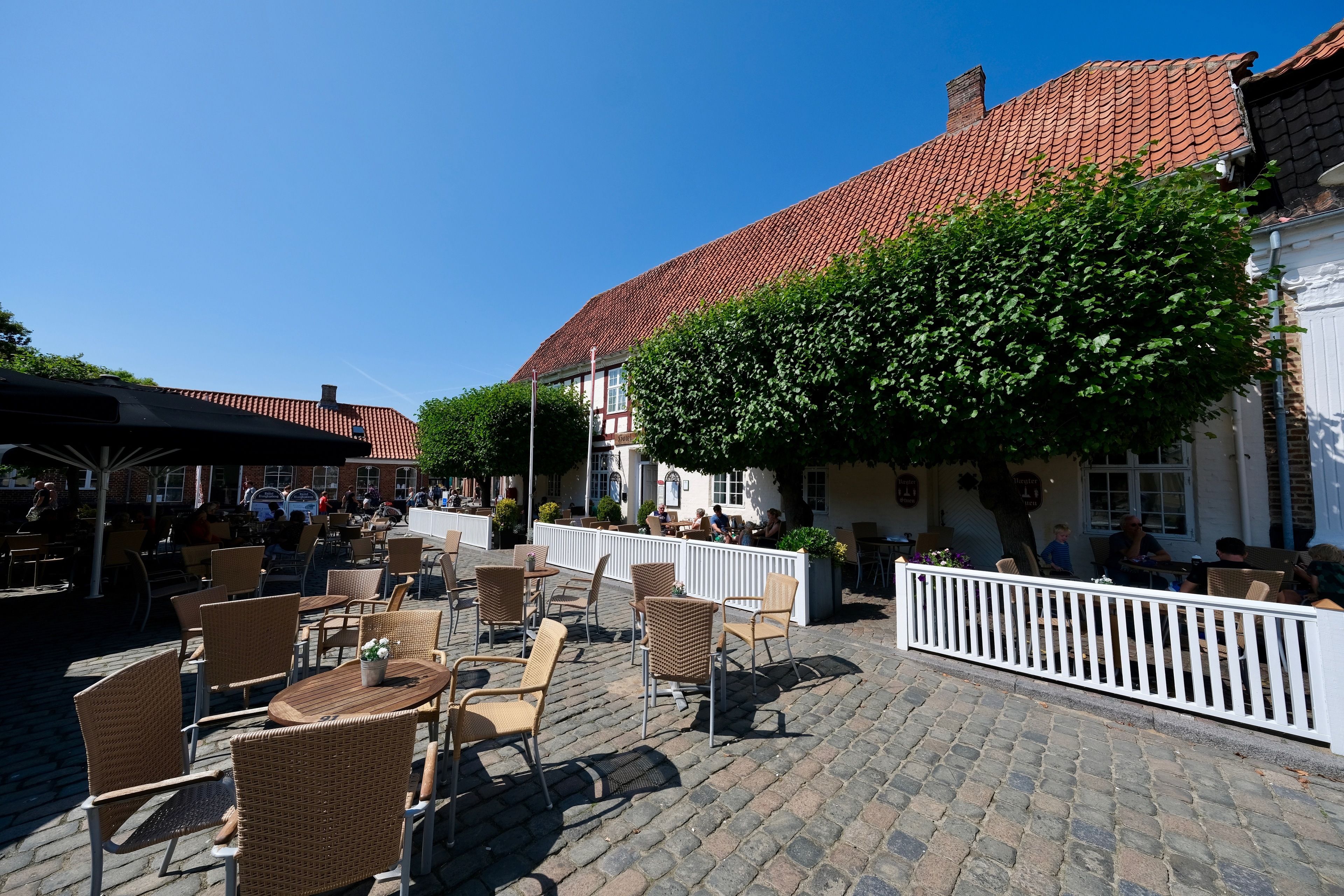 Een terras met rieten stoelen en tafels, een geplaveid terras en een gebouw met een rood pannendak op de achtergrond.