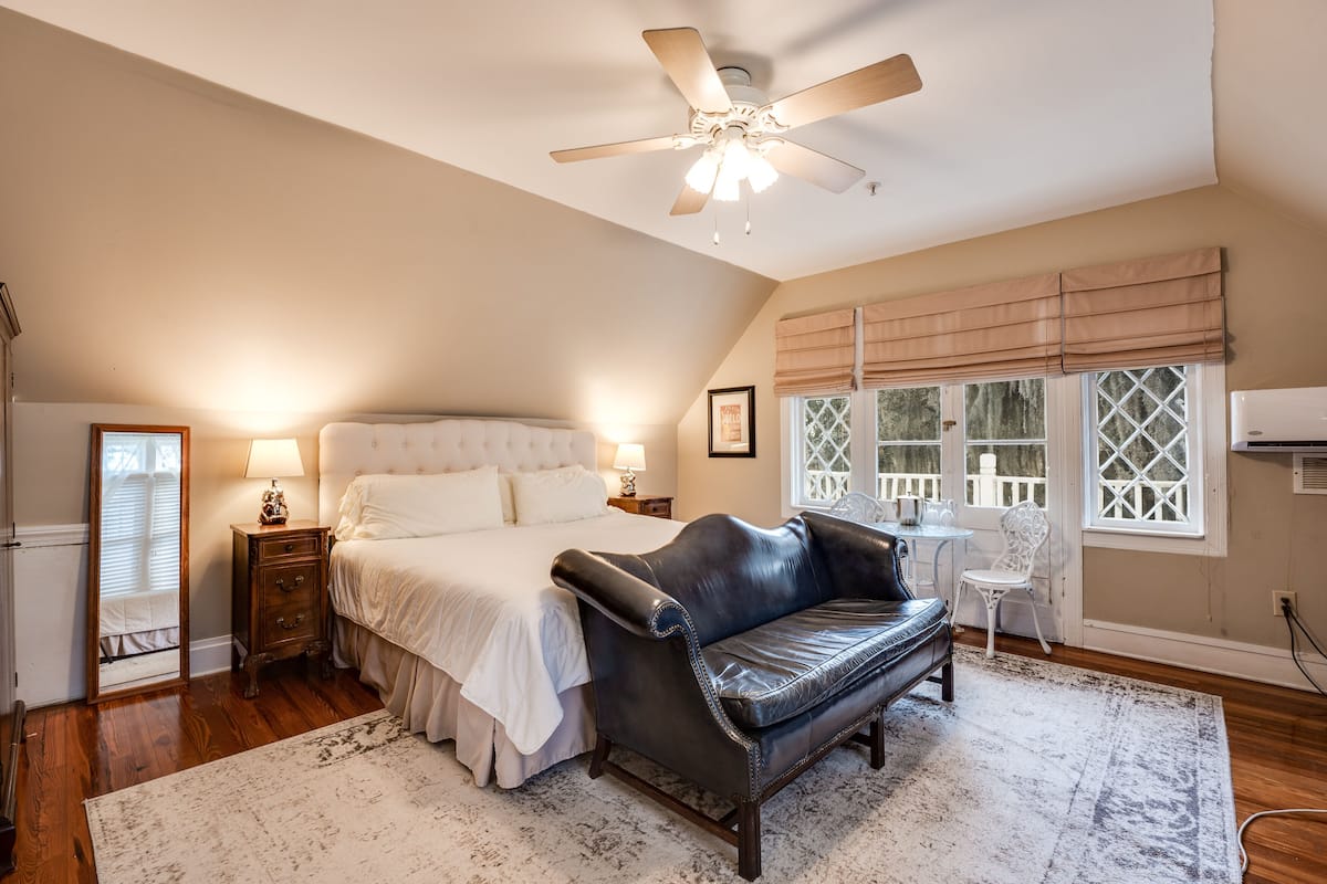 A bedroom with a bed, a leather sofa, a ceiling fan, a window with blinds, and a small table with a chair.