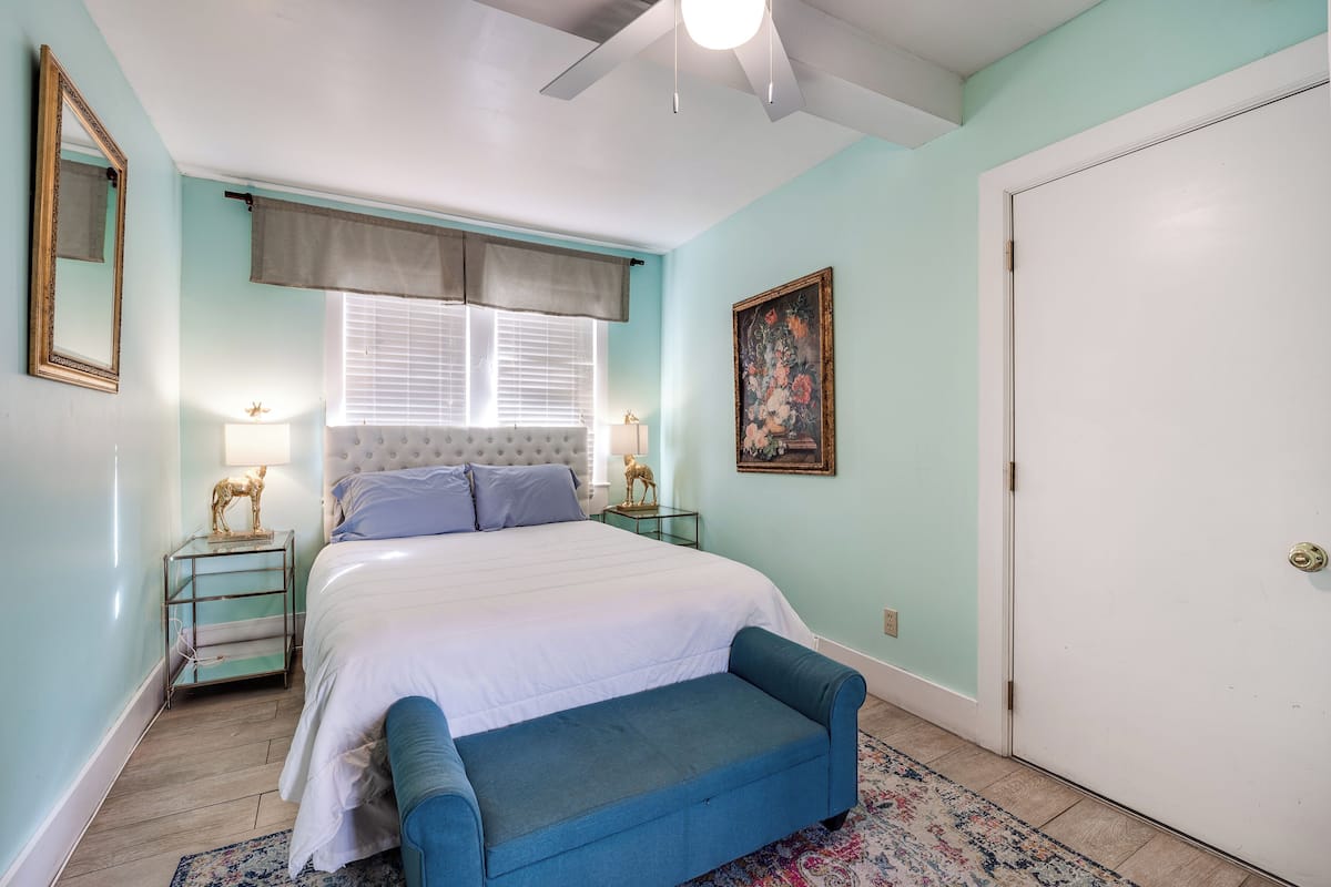 A bedroom with a bed, bedside tables, a bench, a door, and a window with blinds.