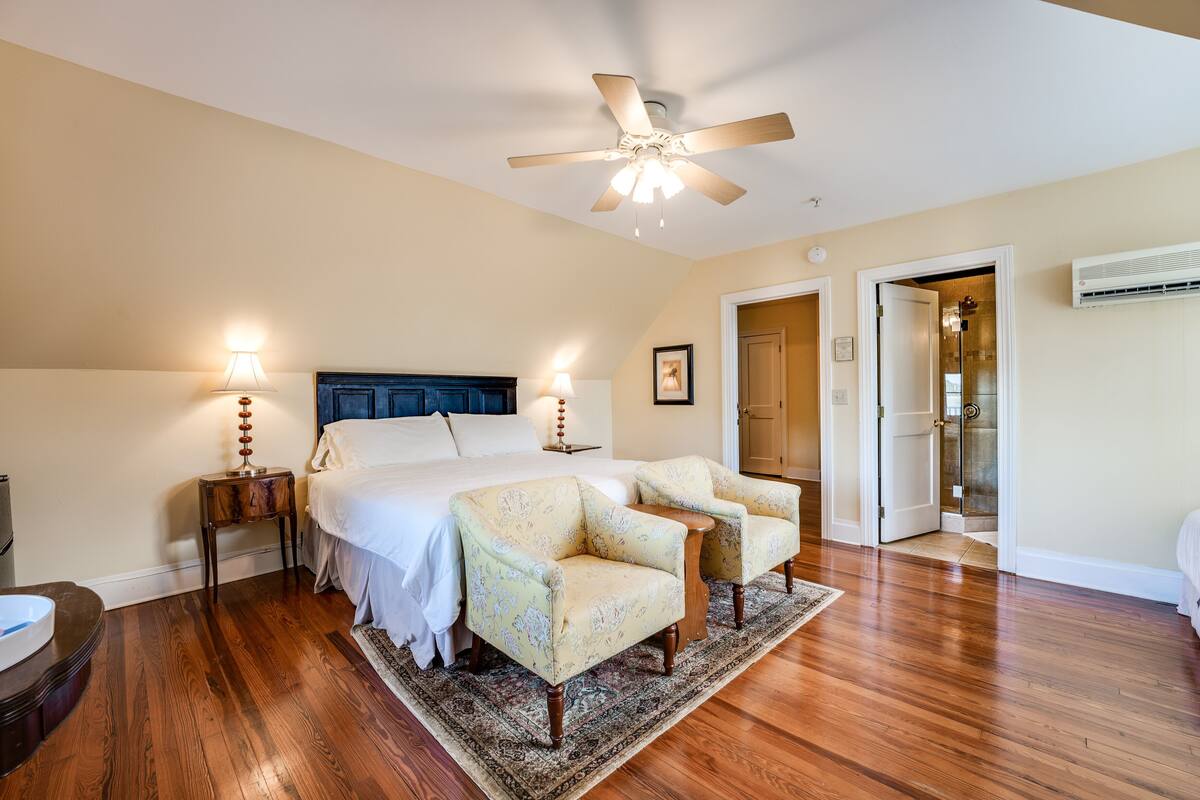 A bedroom with a bed, two armchairs, a nightstand, a ceiling fan, and a door leading to another room.