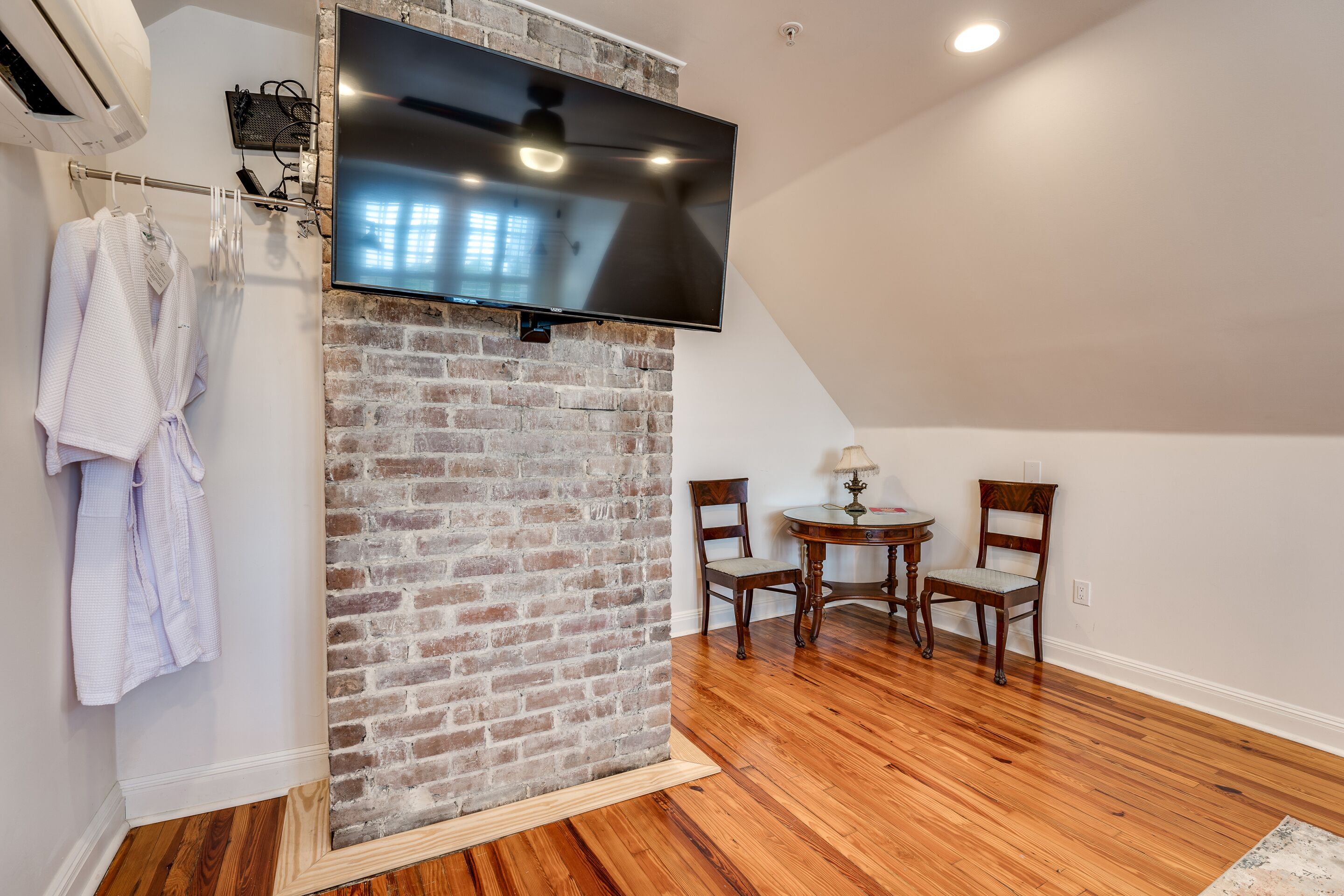 A room with a brick wall, a flat-screen TV mounted above it, wooden flooring, and two wooden chairs.