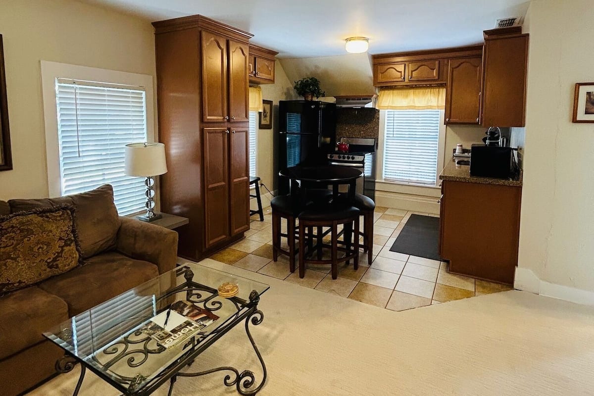 A living room with a sofa, coffee table, and a dining area with chairs and a table. There is a kitchen area with cabinets and appliances.