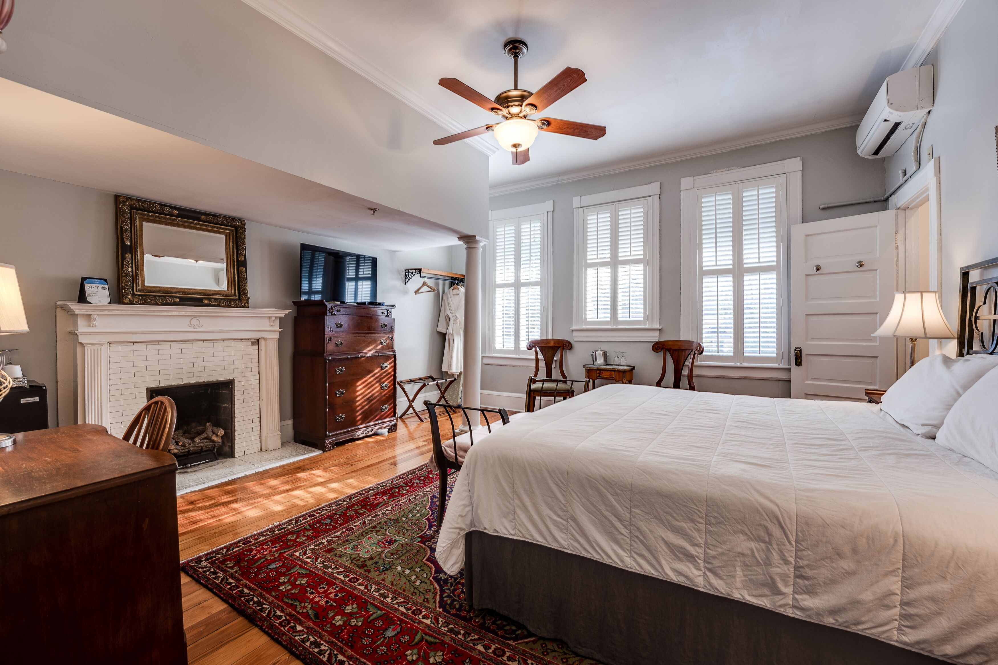 A bedroom with a large bed, a fireplace, a ceiling fan, and a view of the outside through large windows.