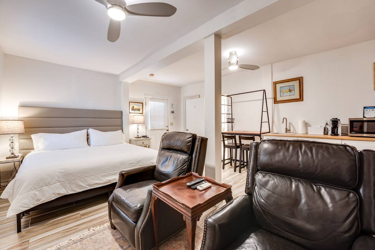A hotel room with a large bed, two leather chairs, a wooden side table, a desk with a chair, a microwave, and a coffee maker.