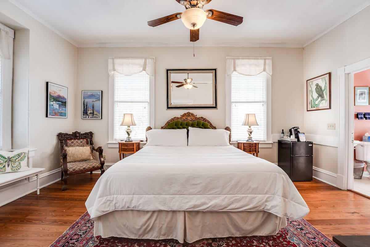 A spacious bedroom with a large bed, two nightstands, a chair, a ceiling fan, and a refrigerator.