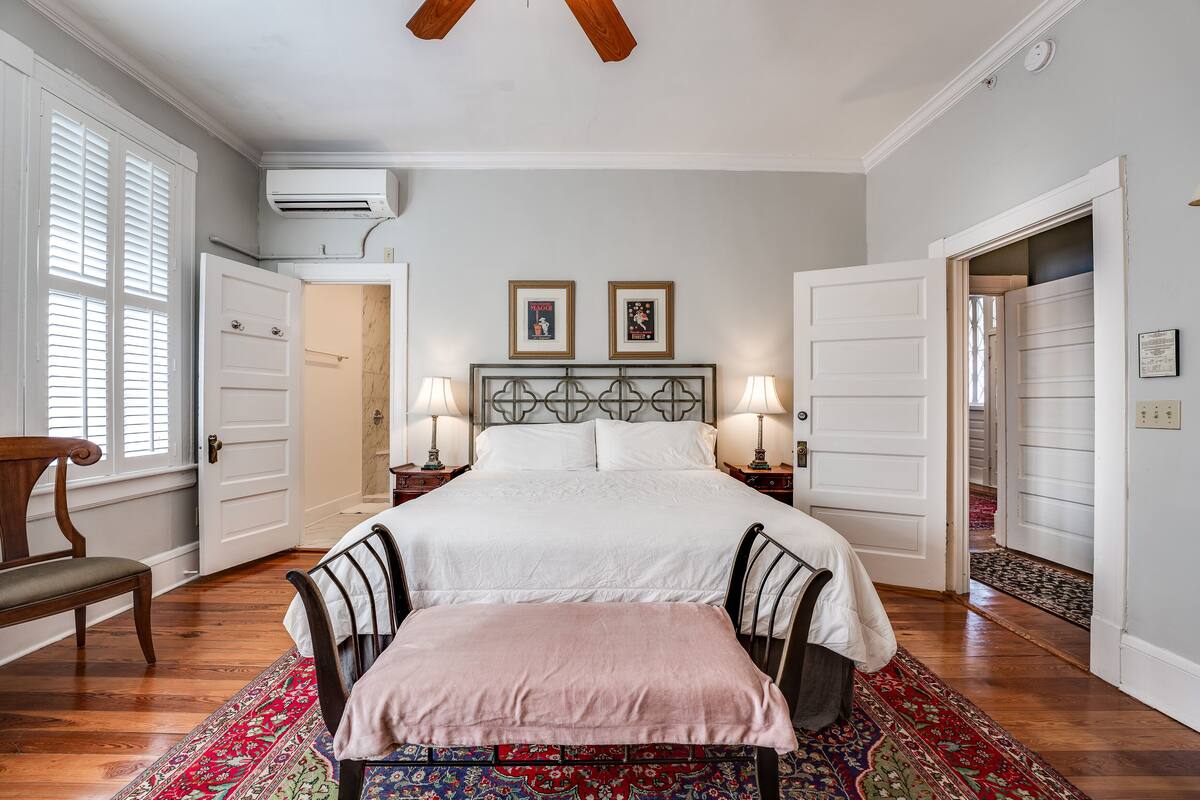 A bedroom with a large bed, two bedside tables with lamps, a bench, a wooden chair, a patterned rug, and a ceiling fan.