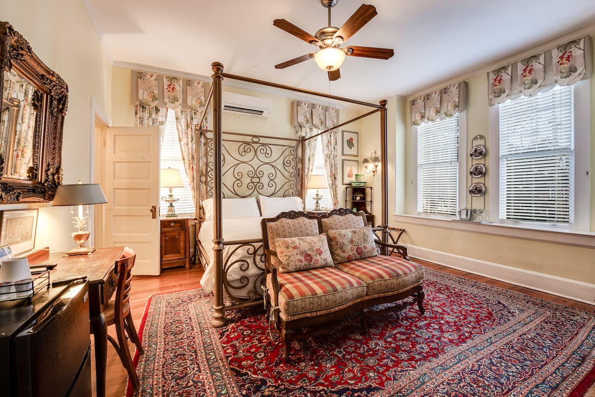 A four-poster bed with a canopy, a wooden desk with a lamp, a red and blue patterned rug, and a view of the outside through a window with blinds.