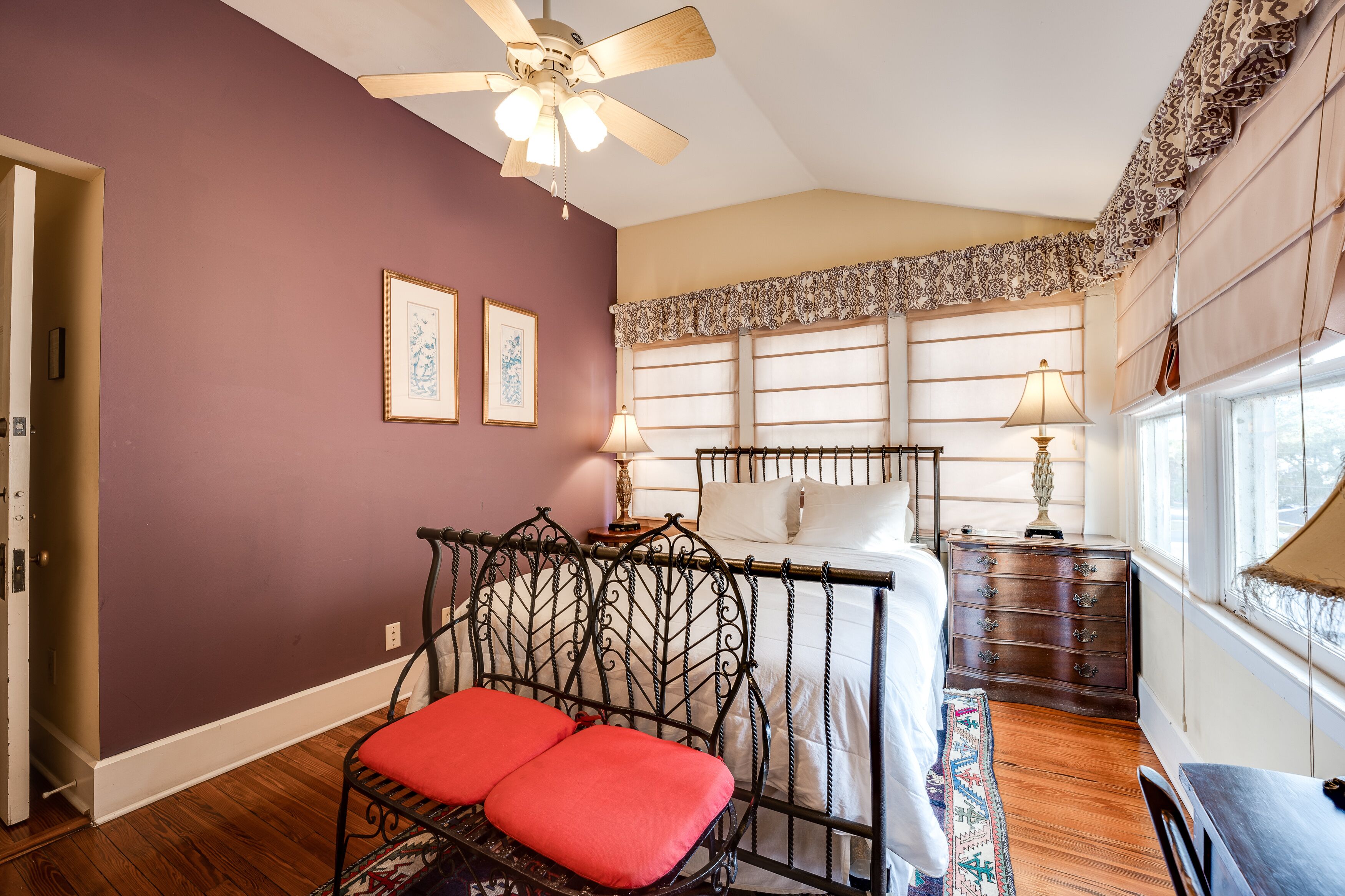 A bedroom with a bed, a chair with red cushions, a ceiling fan, a nightstand with a lamp, and a window with curtains.