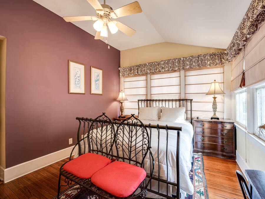 A bedroom with a bed, a chair with red cushions, a ceiling fan, a nightstand with a lamp, and a window with curtains.