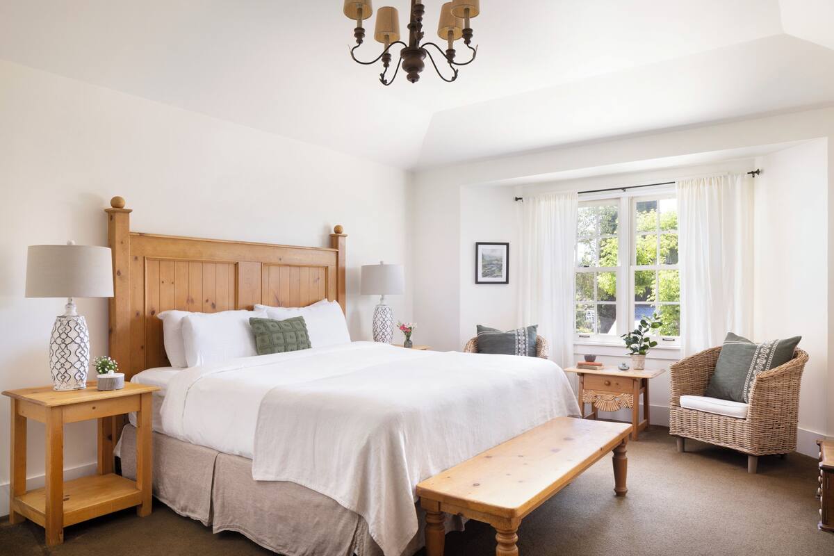 Un dormitorio con una cama de madera, dos mesitas de noche con lámparas, un banco, una silla de mimbre y una ventana con cortinas.