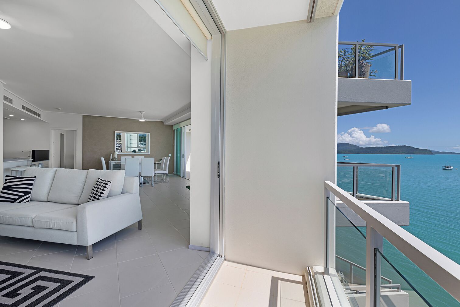 Un salón moderno con un sofá blanco, un comedor y un balcón con vistas a un mar azul y despejado con barcos.