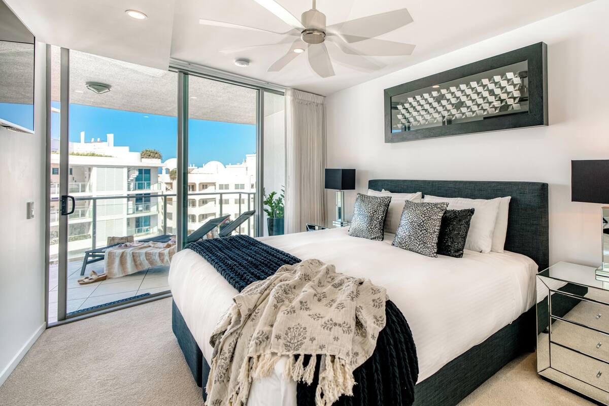 A modern hotel room with a large bed, a ceiling fan, a balcony with a view of the city, and a decorative wall panel.
