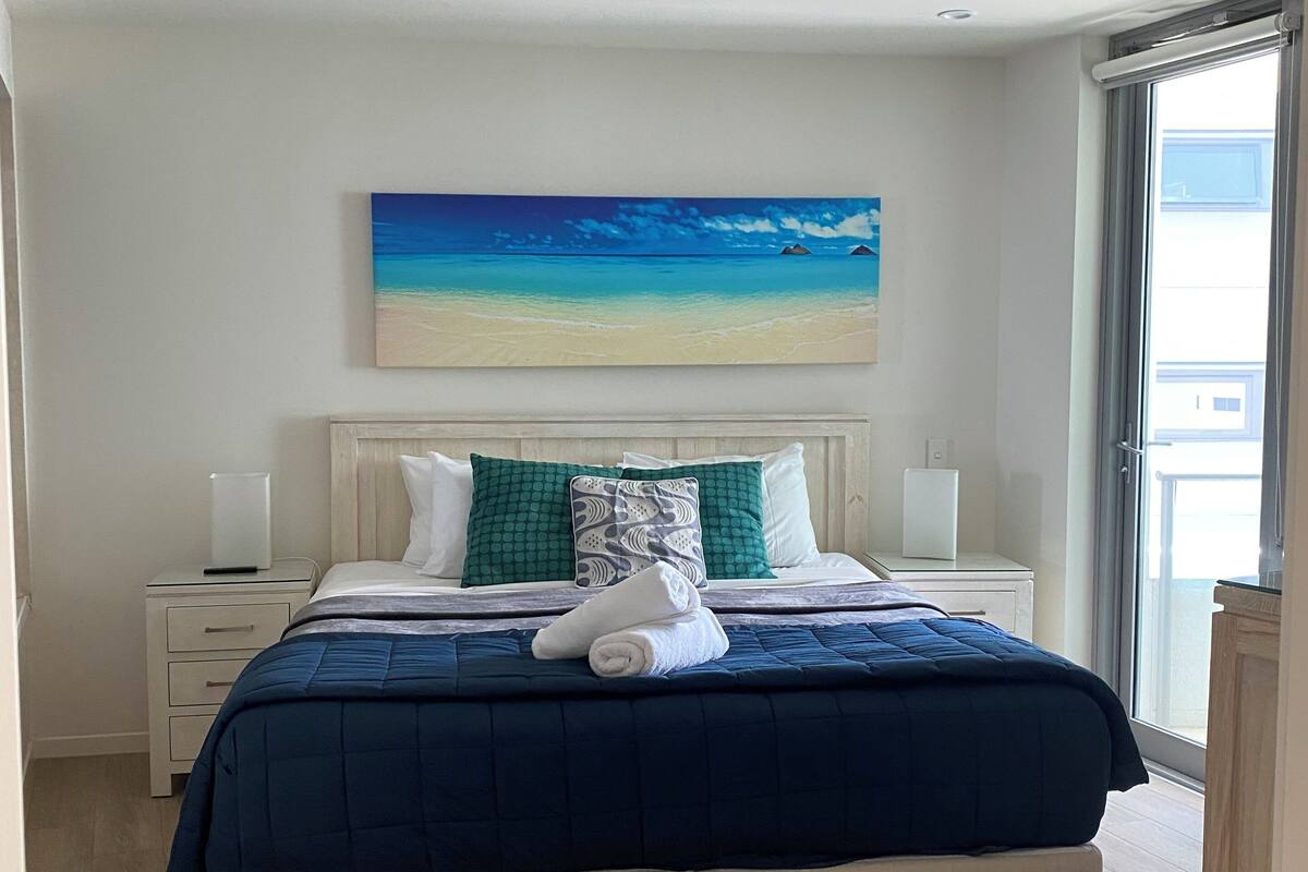 A bedroom with a bed, a painting of a beach, a nightstand, and a window.