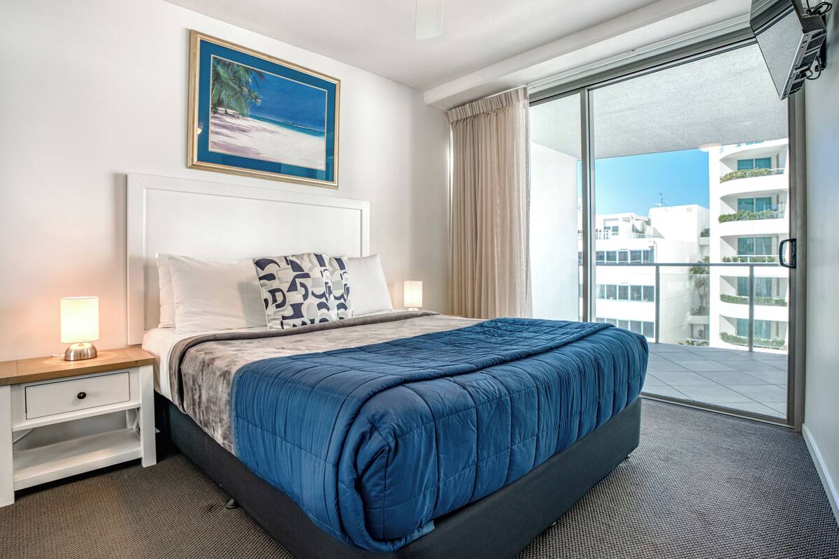 A hotel room with a large bed, a nightstand with a lamp, a painting of a beach scene, and a view of a building through a sliding glass door.