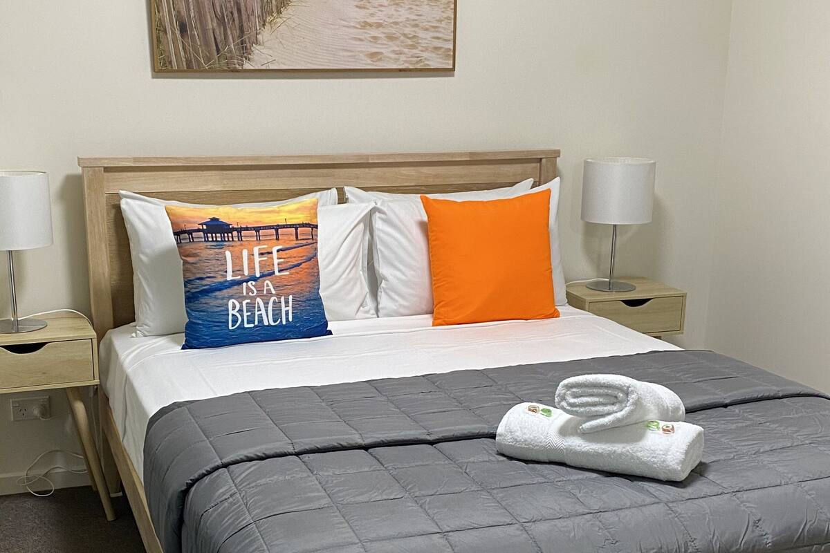 A neatly made bed with a grey quilt, pillows with a beach-themed design, and a folded towel on the bed.