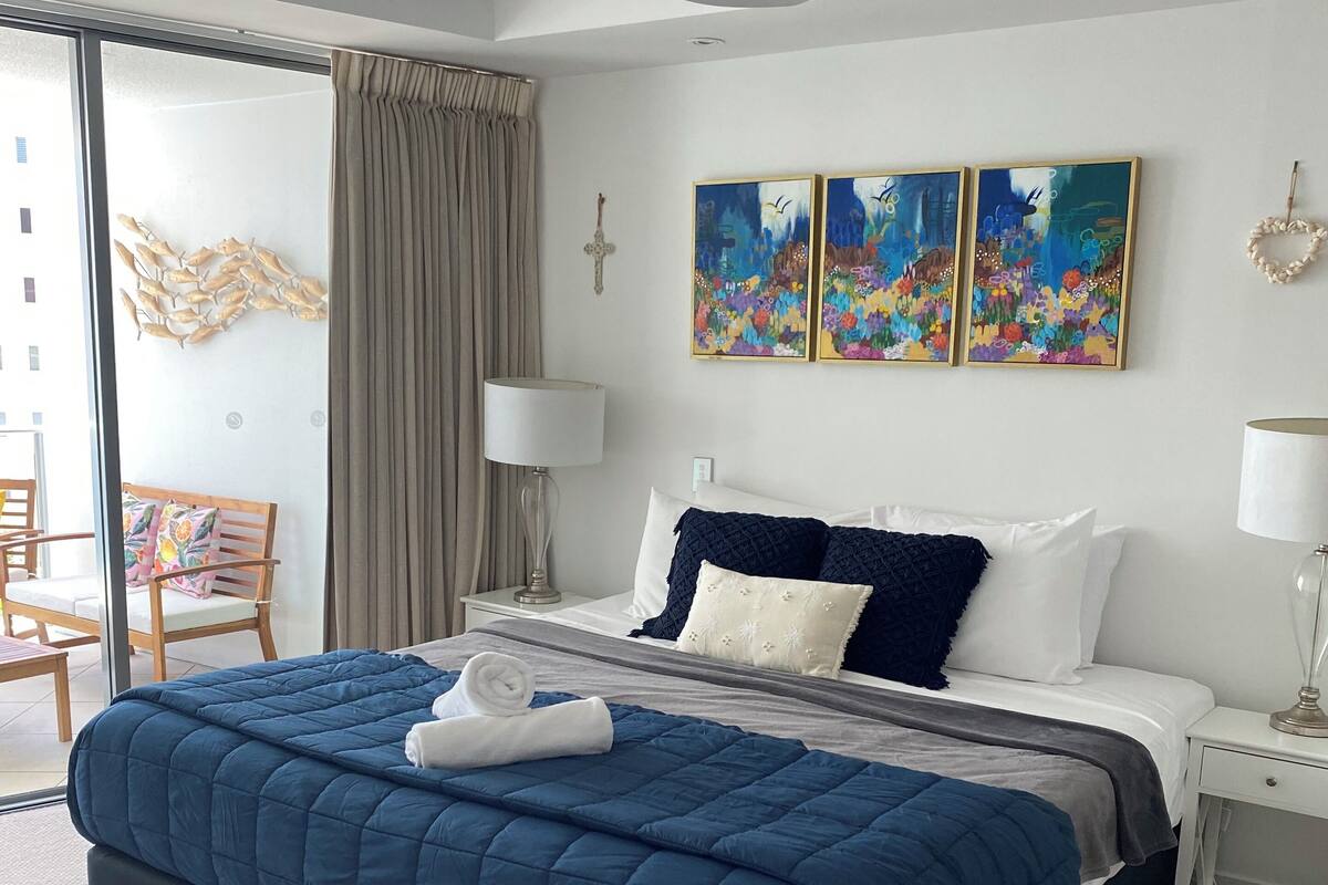 A hotel room with a large bed, a ceiling fan, a cross on the wall, and two paintings.