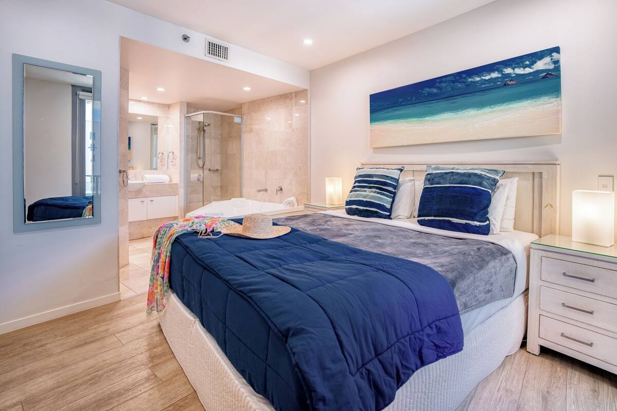 A modern bedroom with a large bed, a nightstand, a bedside table, a mirror, a bathroom with a shower, and a painting of a beach scene.